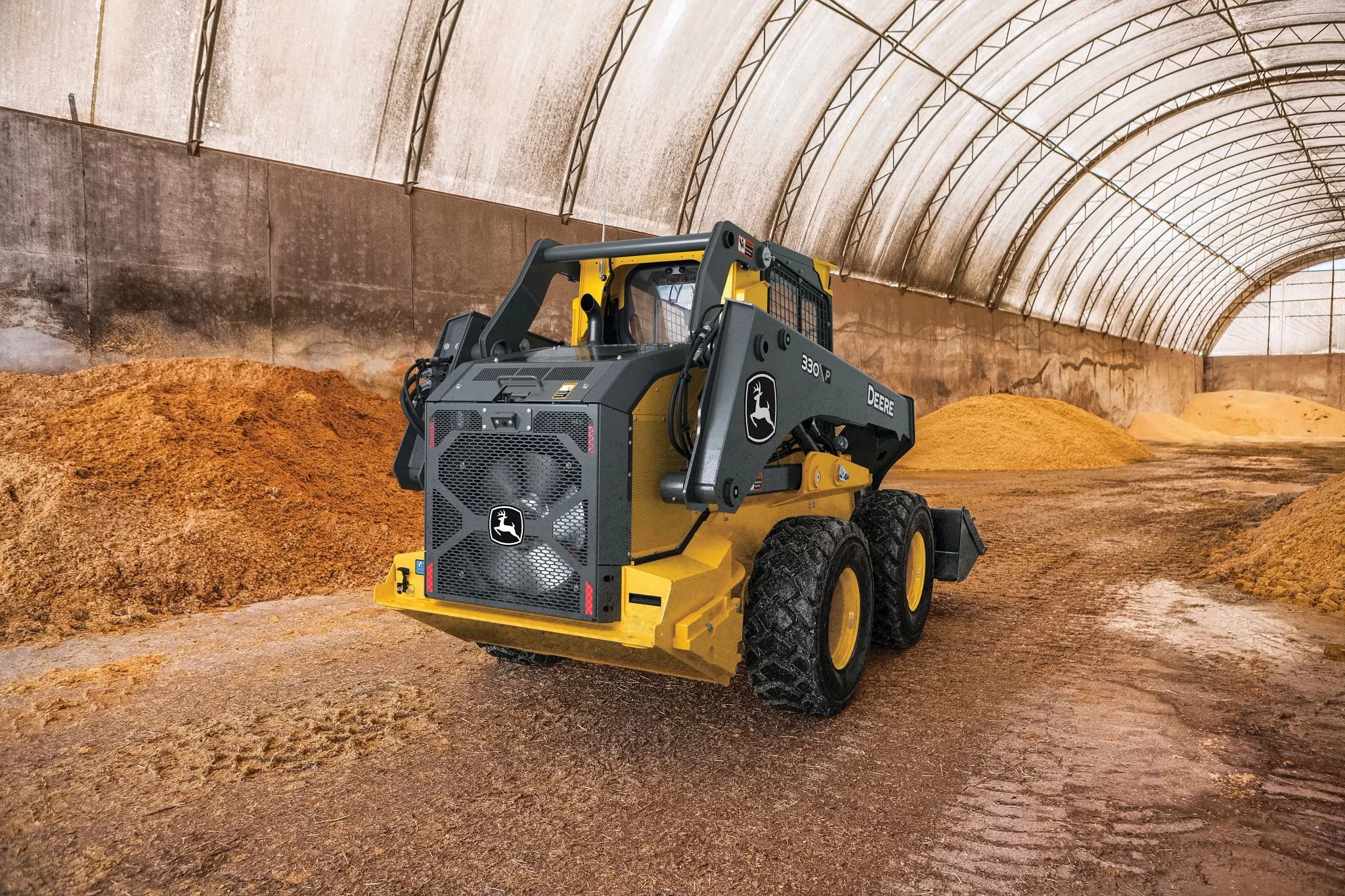 330 P skid steer loader in black and yellow is parked inside a large arched storage building with a translucent roof.