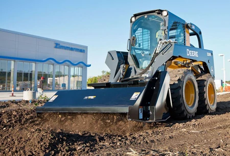 Skid steer loader with a wide front grading attachment leveling soil at a construction site.