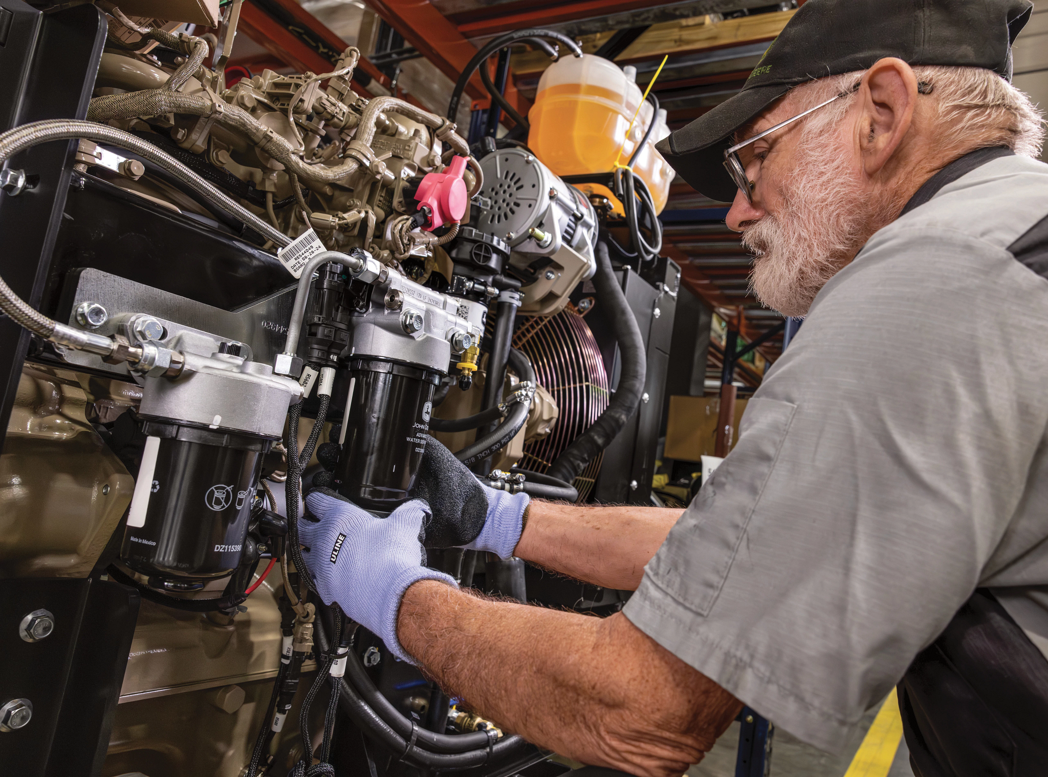 An older male technician wearing gloves and a cap works on a large industrial engine, installing a mounted filter assembly amid hoses, wiring, and metal components in a workshop