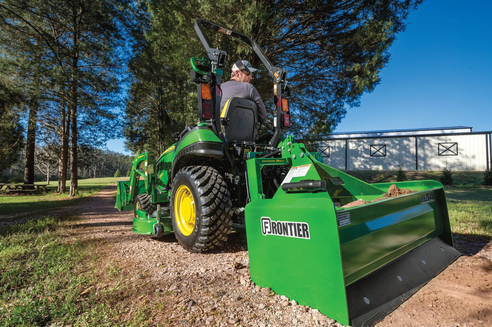 Green tractor equipped with a rear-mounted box blade leveling gravel on a path near trees and a light-colored building, with a person seated in the driverâ€™s position