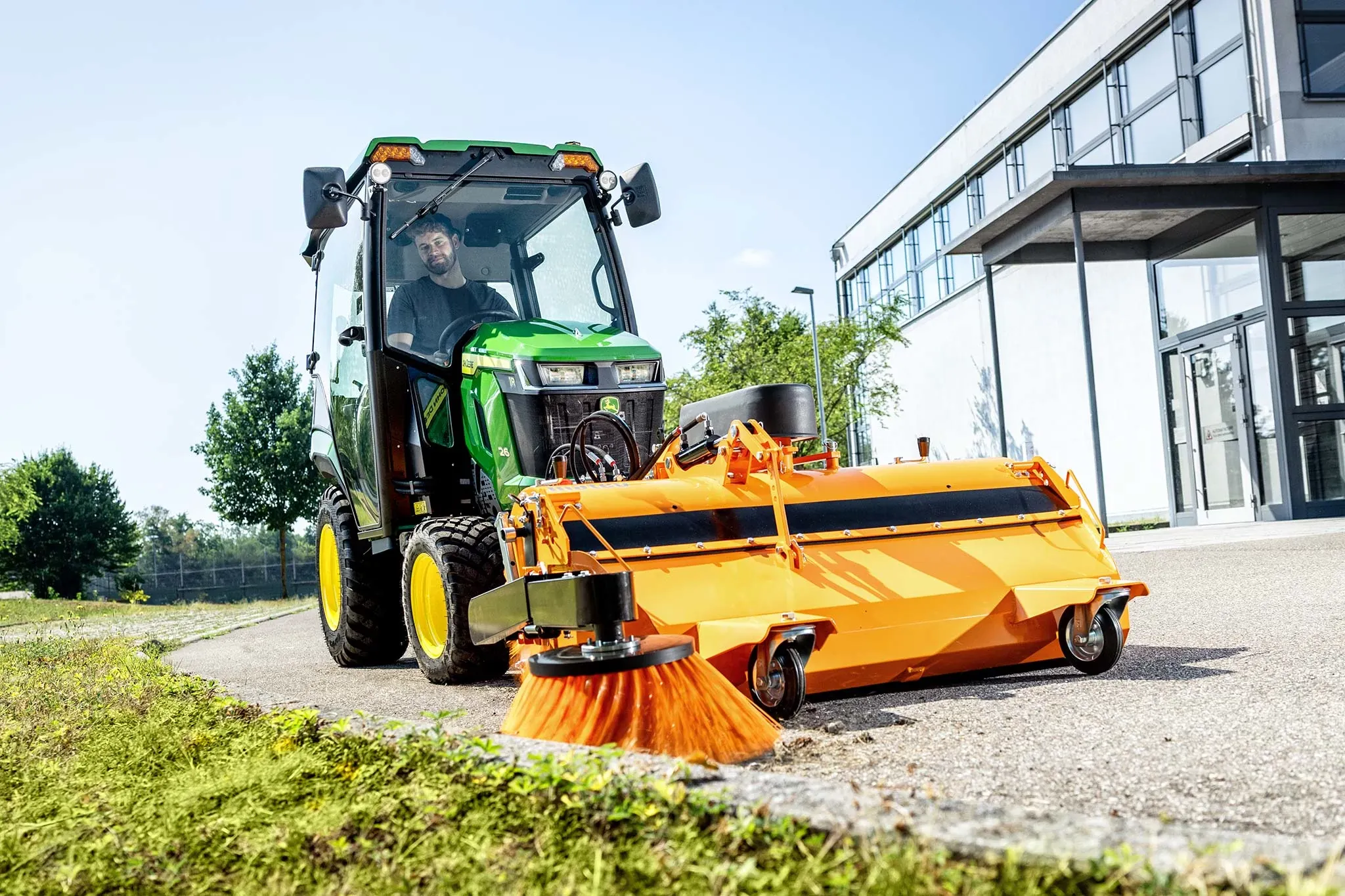 John Deere compact tractor with enclosed cab and front-mounted rotary broom cleaning a paved area