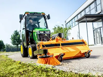 John Deere compact tractor with enclosed cab and front-mounted rotary broom cleaning a paved area
