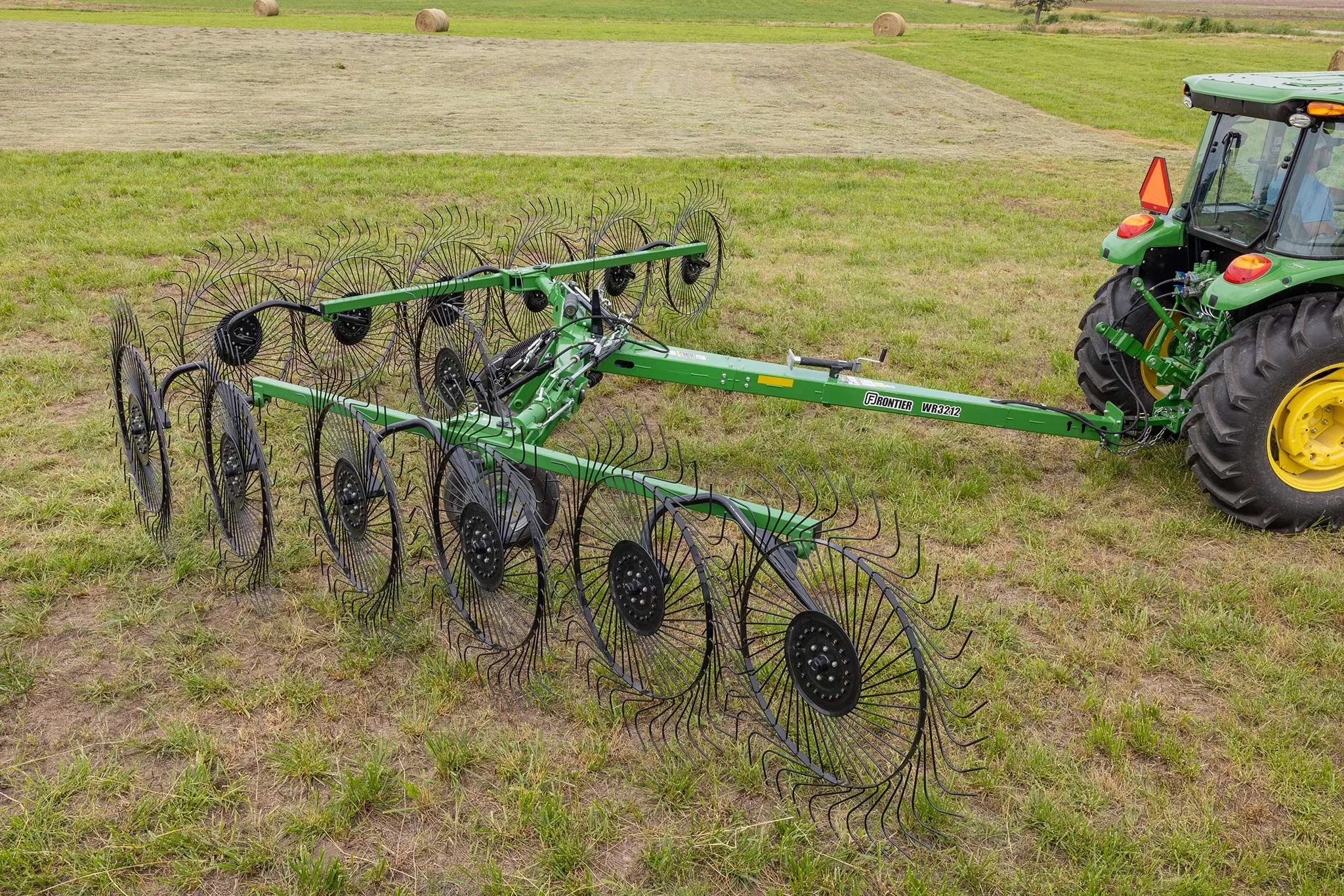 Side right view of a Frontier WR3212 wheel rake resting on grass, with the back end of a 5E tractor visible on the right side of the frame
