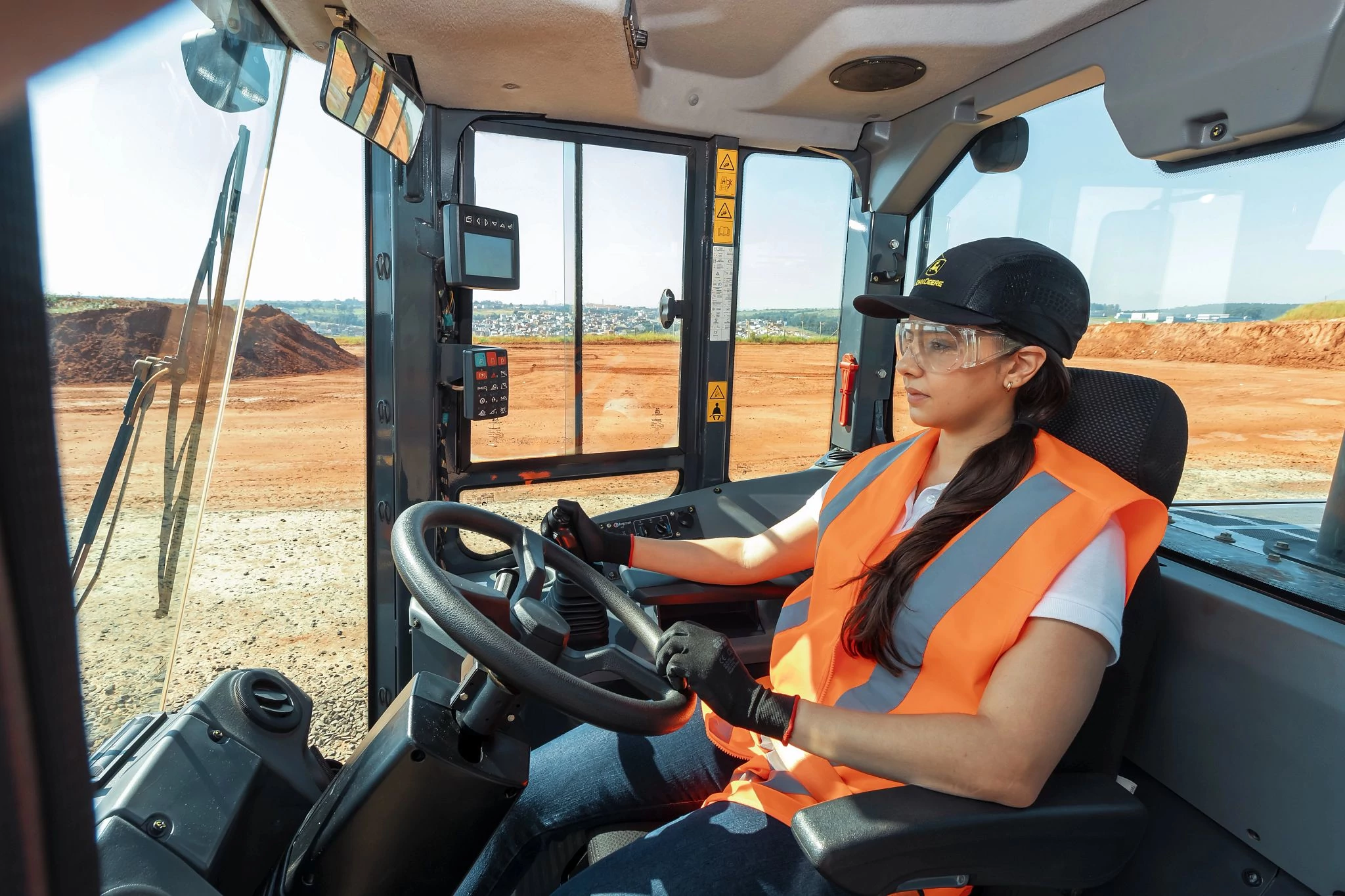 A person wearing a hard hat, safety glasses, gloves, and a high-visibility orange vest sits inside the cab of a heavy construction vehicle, holding the steering wheel
