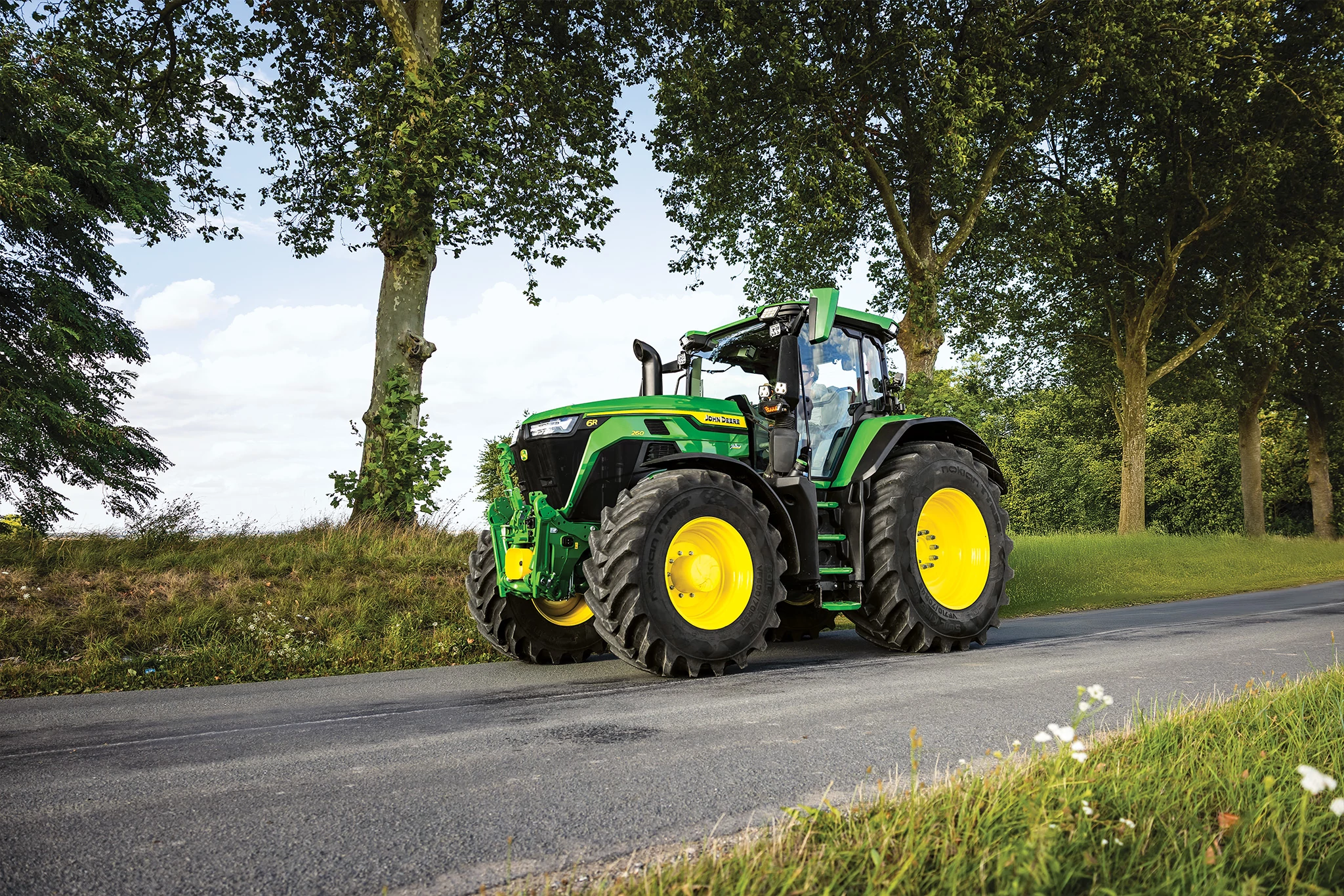 Side left view of a 6R 260 tractor traveling down a tree-lined country road.