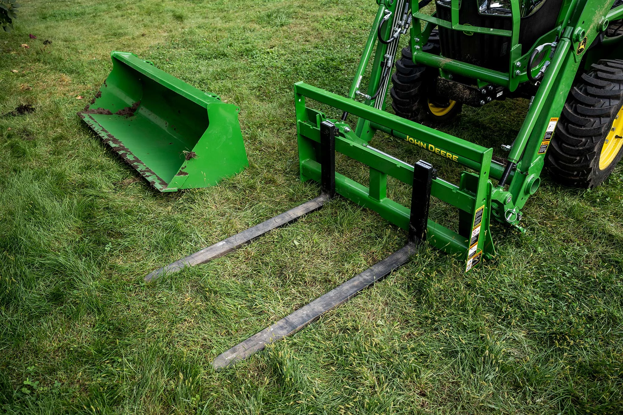 Top left view of an AP11F pallet fork attached to a John Deere compact tractor with a green bucket laying in the grass beside it.