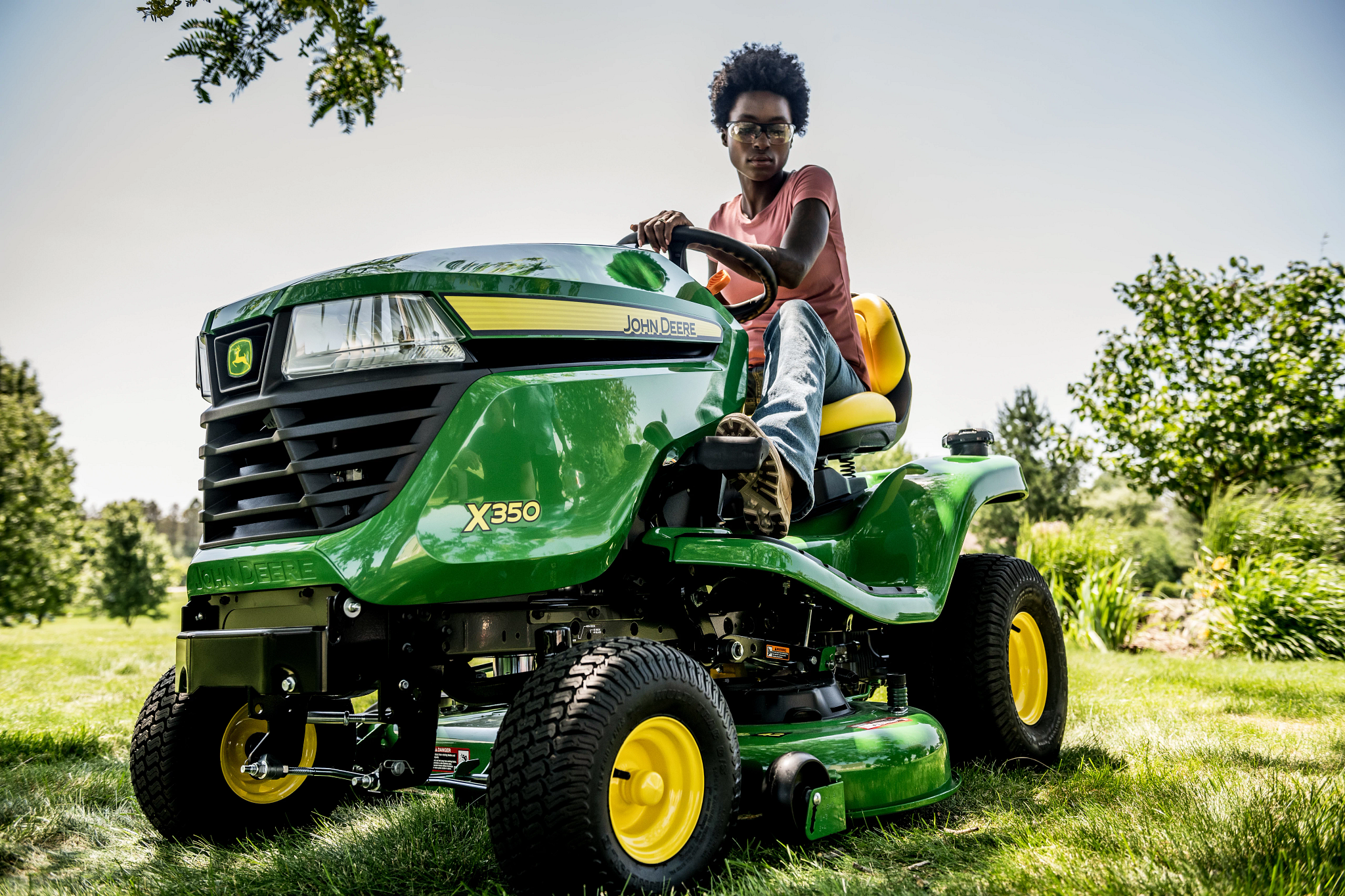 Person seated on a green John Deere X350 riding mower on a grassy lawn, with trees and landscaping visible in the background