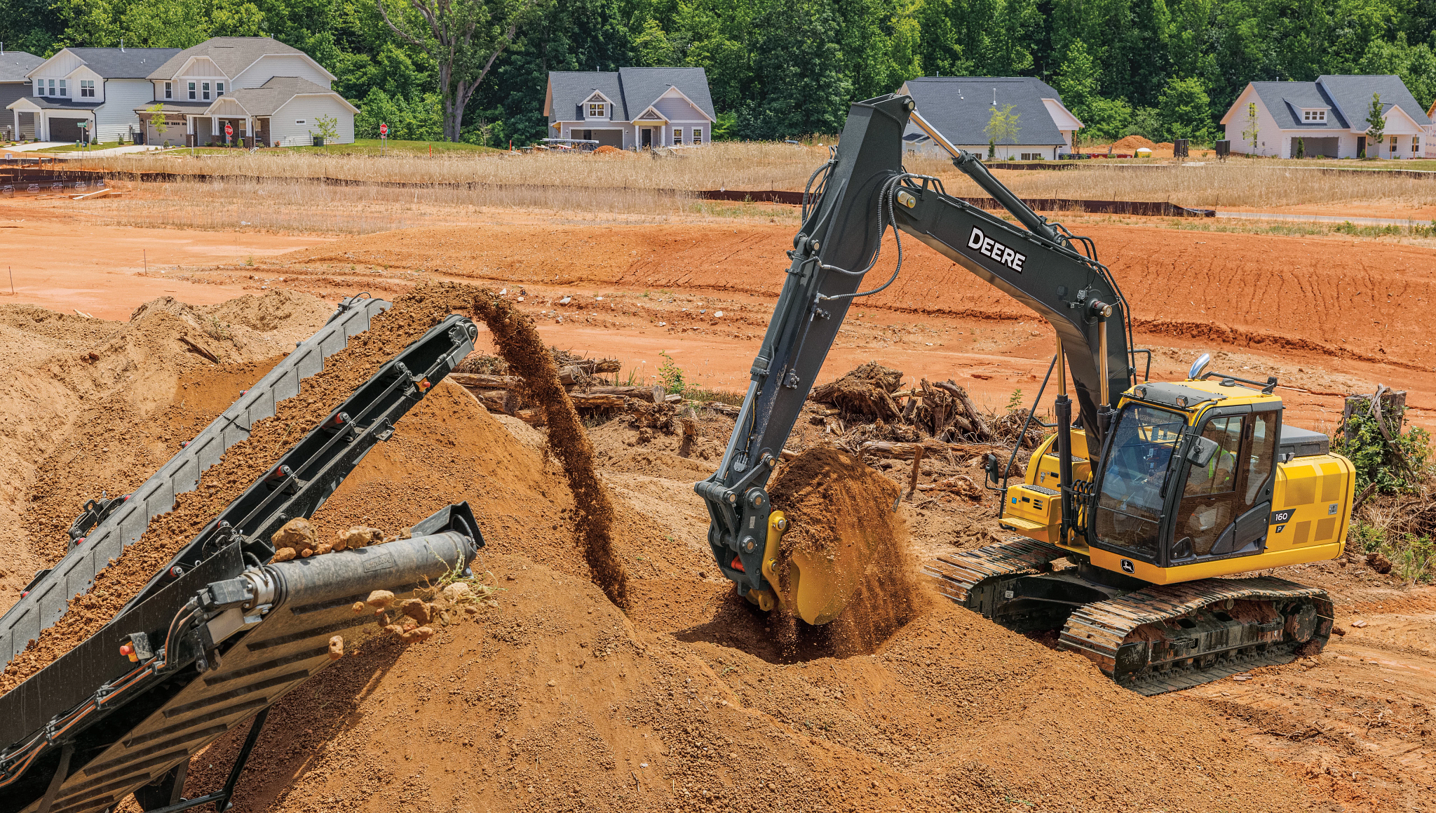 Yellow excavator loading reddish-brown soil into a conveyor-fed screening machine at a construction site, with dirt being poured into a pile while a row of houses and trees appear in the background