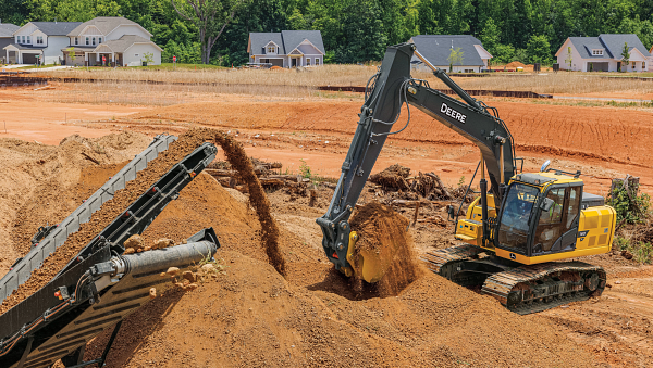 Yellow excavator loading reddish-brown soil into a conveyor-fed screening machine at a construction site, with dirt being poured into a pile while a row of houses and trees appear in the background
