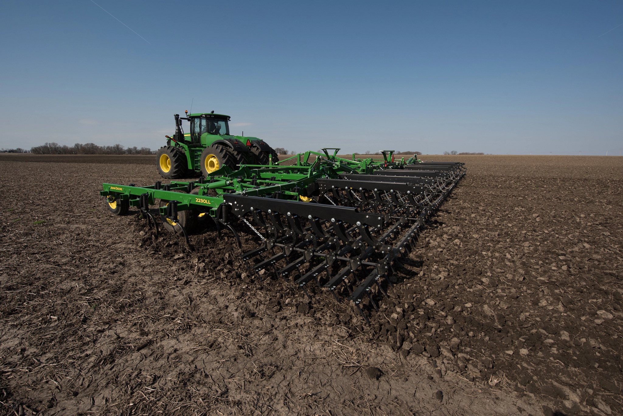 John Deere tractor pulling a wide green field cultivator across a bare field, with multiple rows of shanks and tines working the soil under a clear sky
