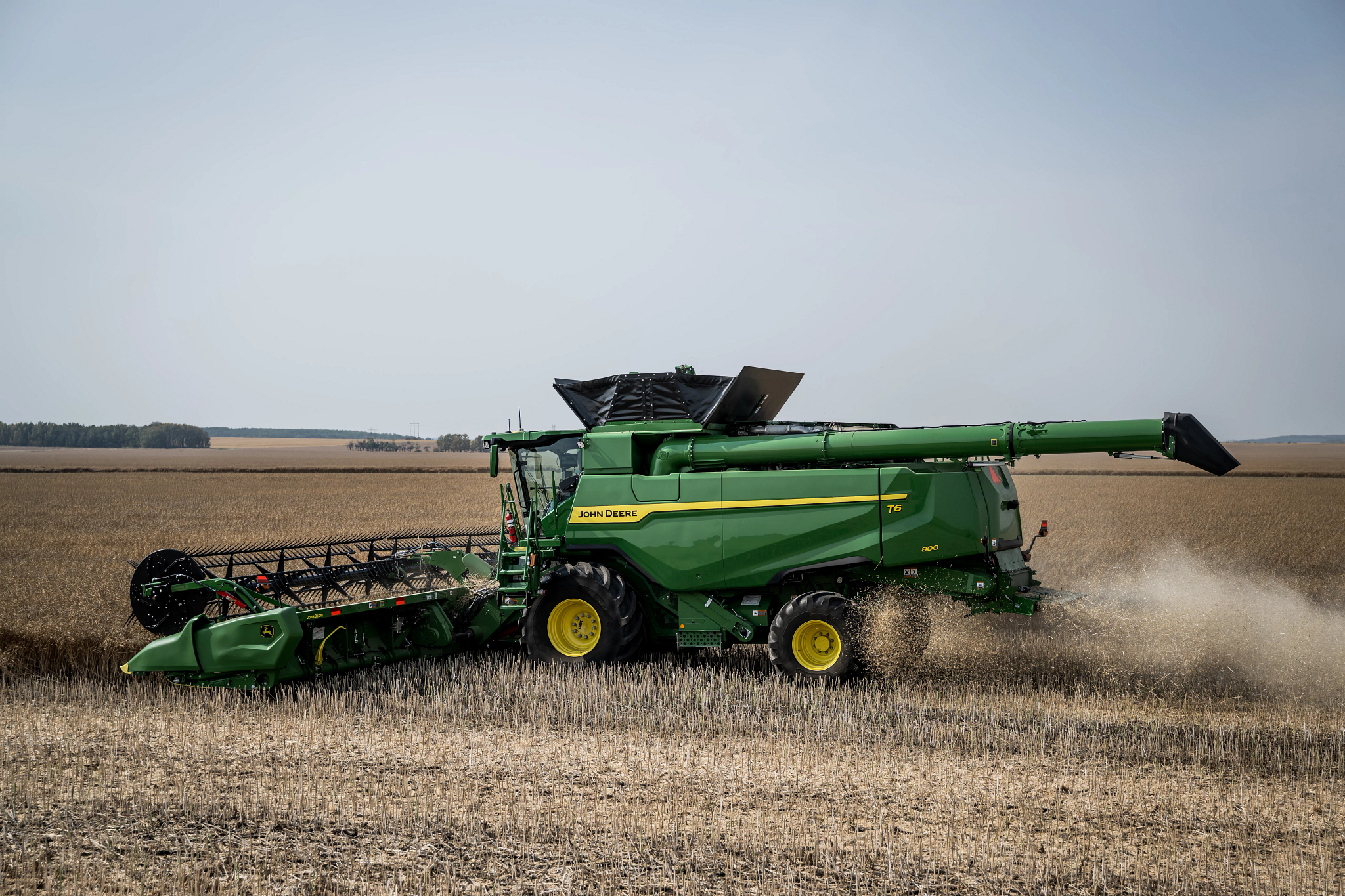 Side view of a combine harvester cutting crops in a wide open field, with the harvesting header engaged at the front, grain unloading auger extended, dust and chaff trailing behind the rear wheels.
