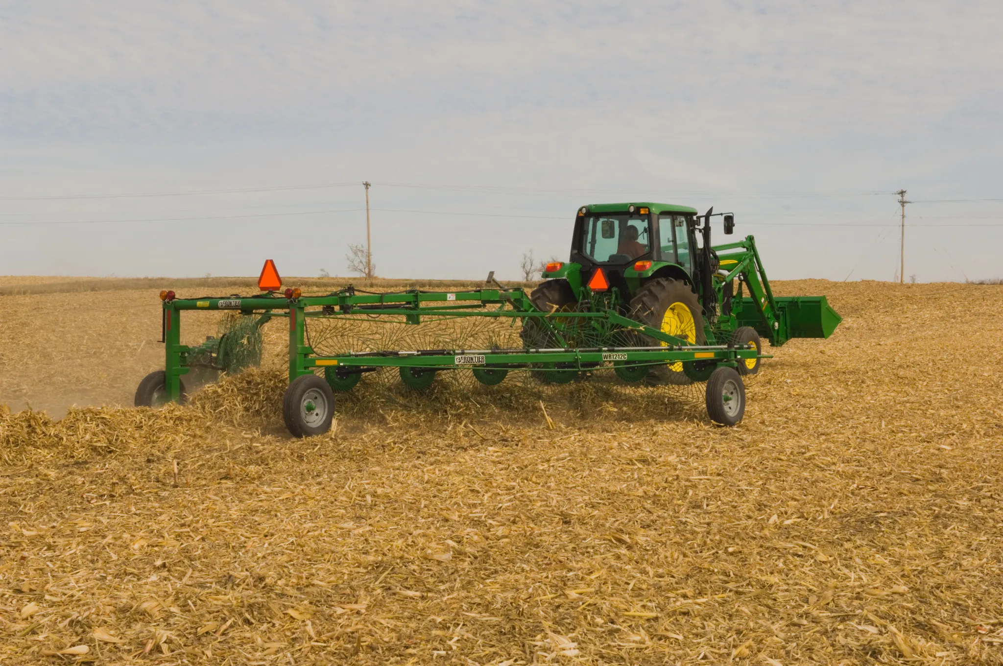 Rear right view of a tractor pulling a WR1212 High-Capacity Wheel Rake through a tan, harvested field.