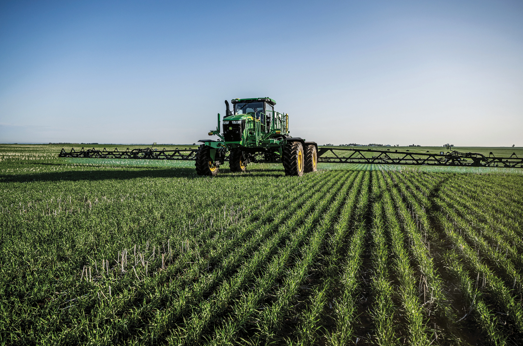 Wide view of a green agricultural sprayer driving through a large, neatly planted field, with long rows of crops stretching into the distance under a clear blue sky