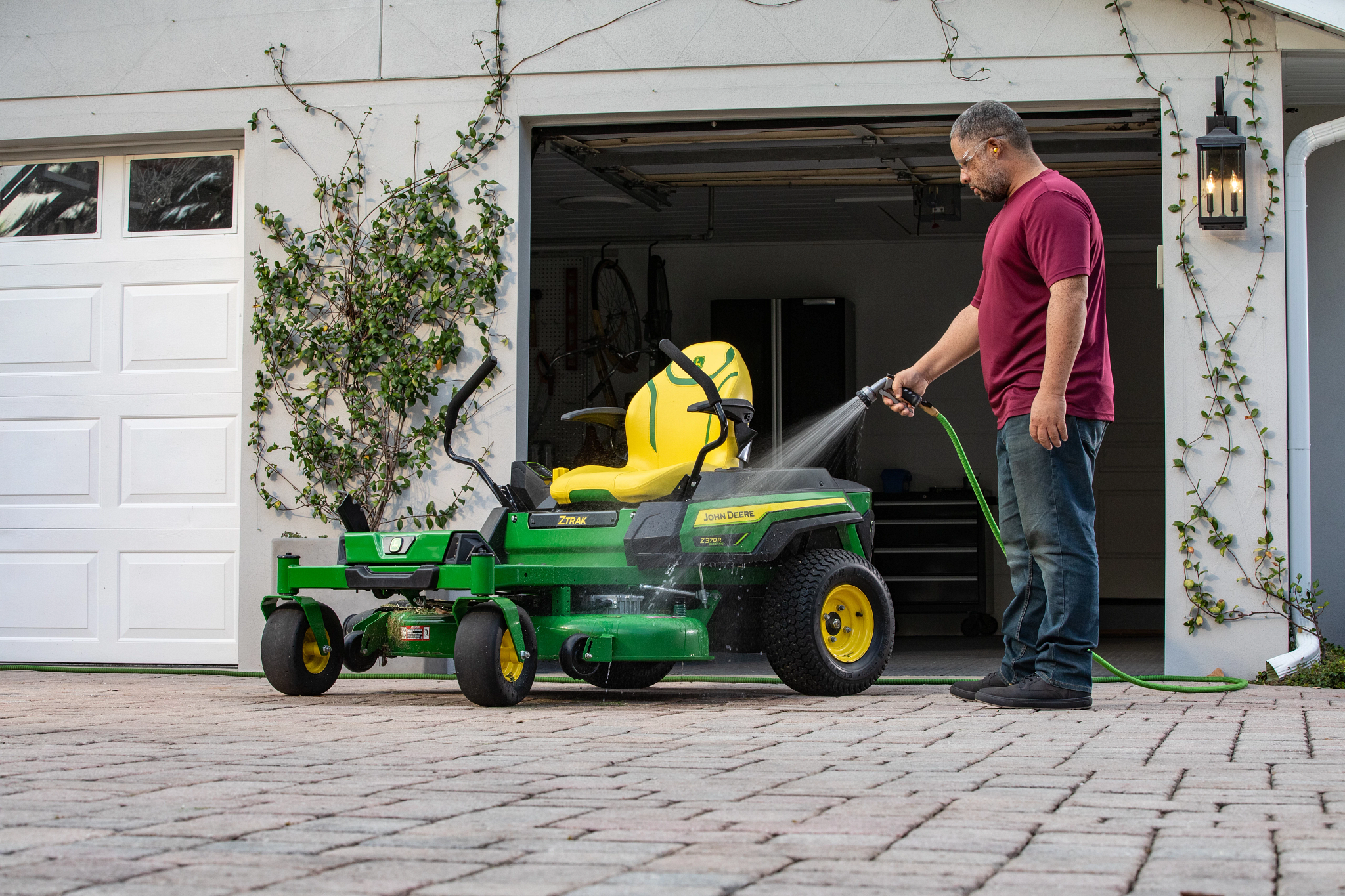 Red-shirted homeowner hosing down a Z370R Electric ZTrak™ Mower on a cobblestone driveway in front of an open-doored garage