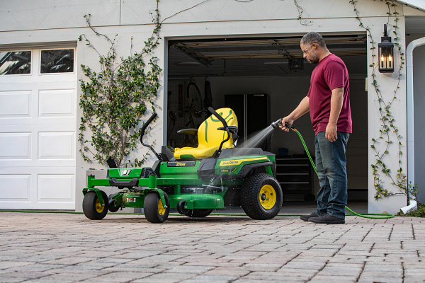Red-shirted homeowner hosing down a Z370R Electric ZTrak™ Mower on a cobblestone driveway in front of an open-doored garage