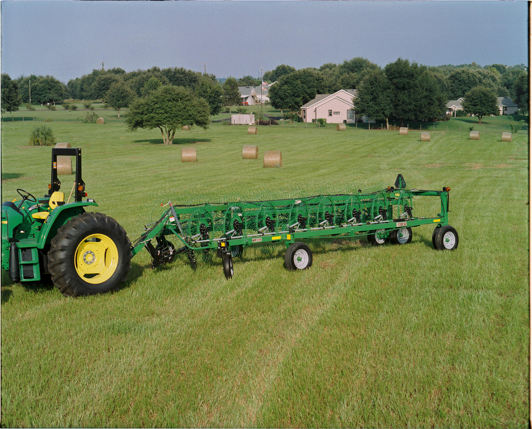 Tractor pulling a long wheel rake across a grassy field with multiple round hay bales scattered in the background