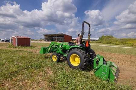 Side view of a green tractor operating a rear-mounted flail mower in a grassy field, with farm buildings and open countryside visible under a partly cloudy sky