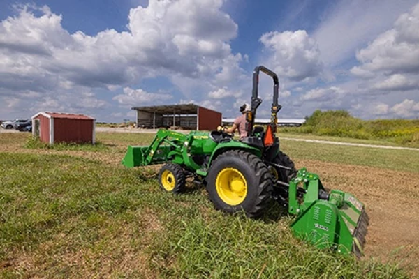 Side view of a green tractor operating a rear-mounted flail mower in a grassy field, with farm buildings and open countryside visible under a partly cloudy sky