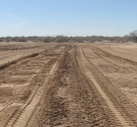 Wide view of an open dirt field with parallel tire tracks running across freshly graded soil under a clear blue sky