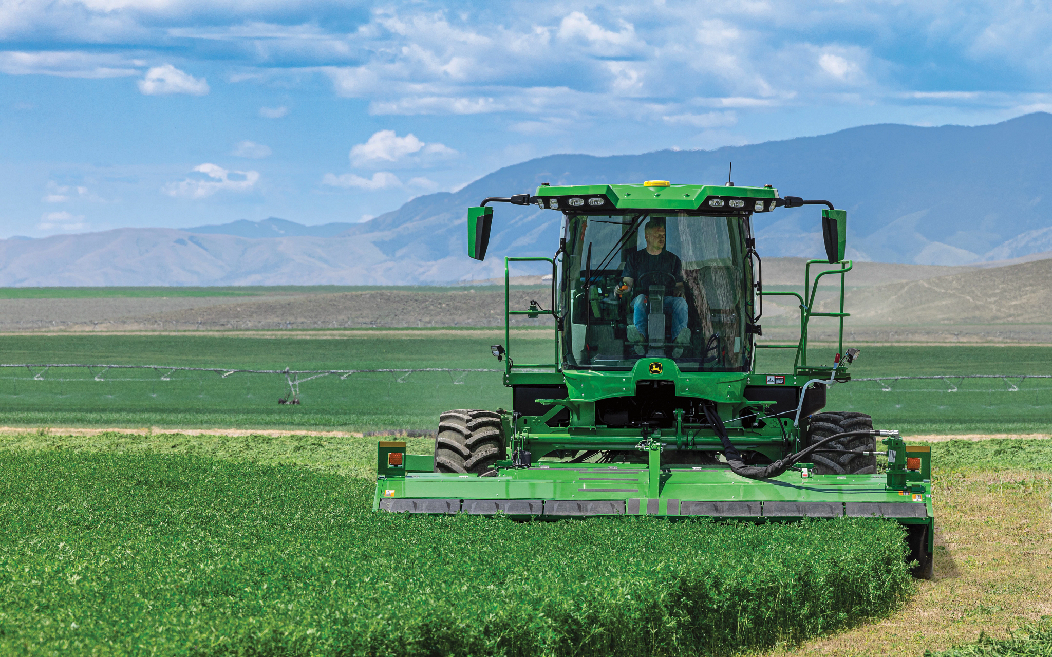 John Deere self‑propelled windrower with enclosed cab and wide mower‑conditioner cutting alfalfa field, operator visible inside, mountains and blue sky in background.