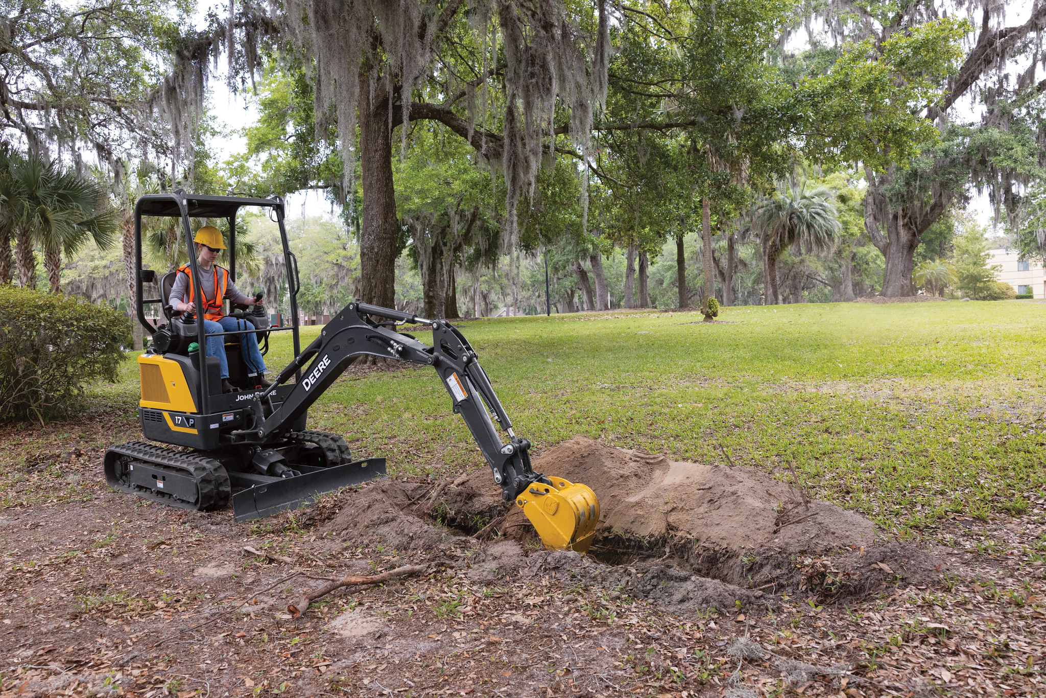 Small excavator digging a shallow trench in a grassy park area, with an operator in a high-visibility vest and hard hat seated inside the cab, surrounded by trees draped with hanging moss