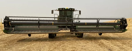 A front view of a wide draper platform attached to a self‑propelled windrower, positioned in a cut wheat field