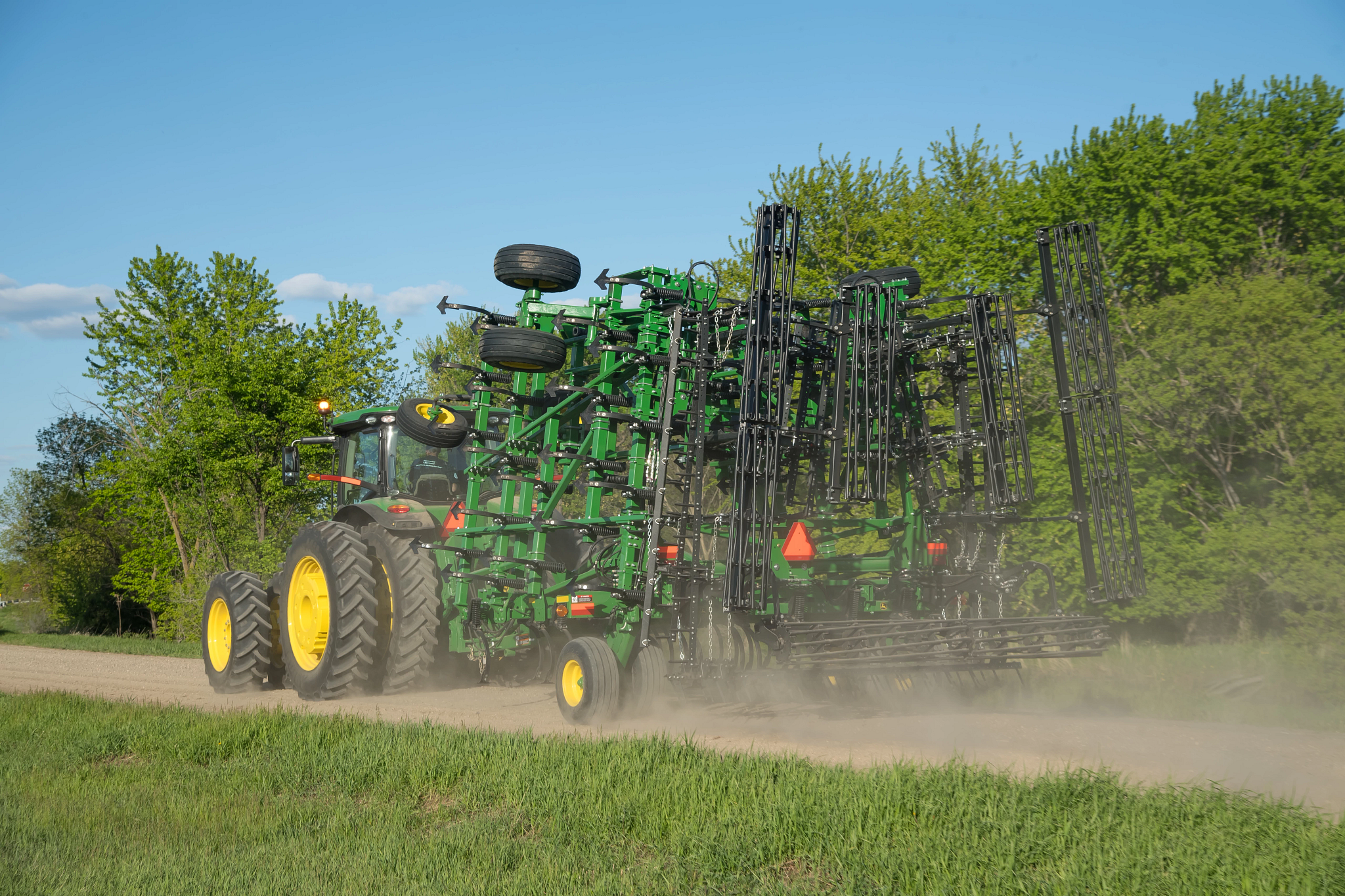 A John Deere tractor travels down a dirt road pulling a folded green field cultivator, kicking up dust with trees and grass in the background