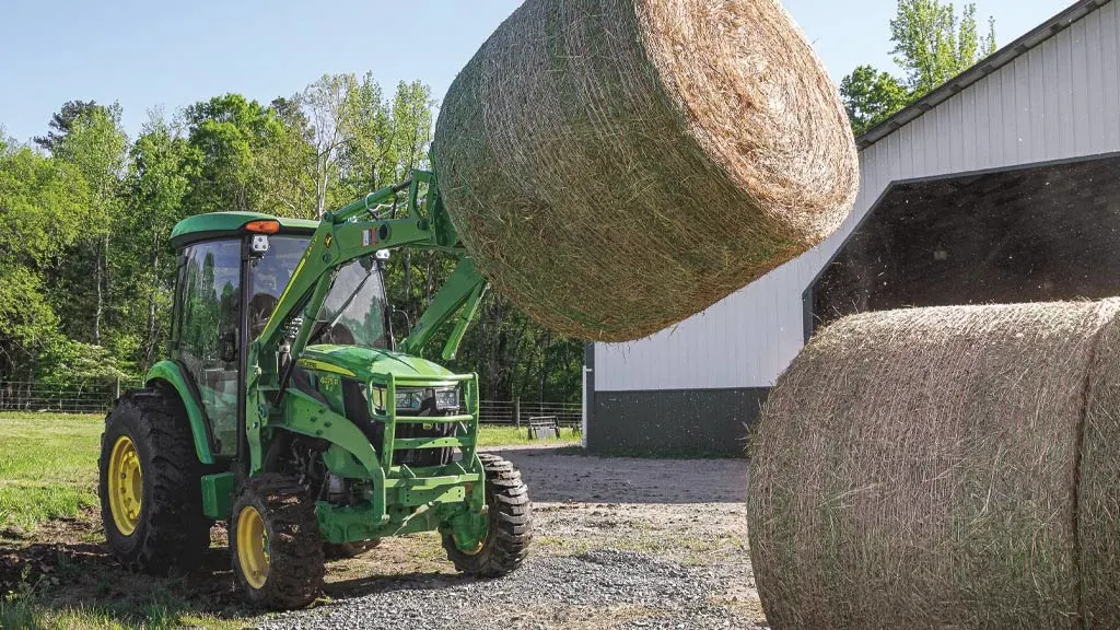 Front right view of a 4R series tractor with front loader lifting a large round hay bale next to another in front of a barn