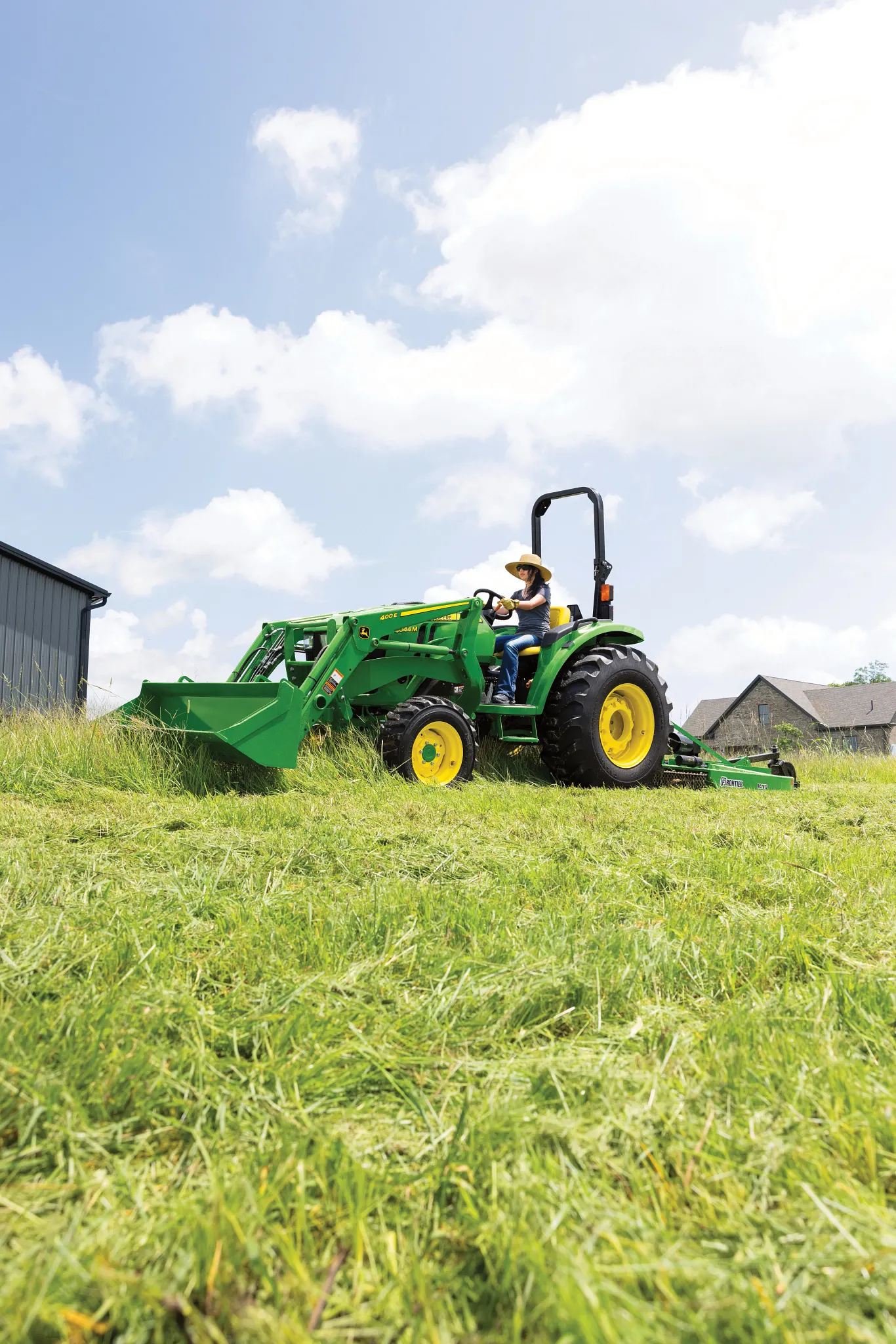 Side left view of a farmer driving a 4044M open operator station tractor across a grassy field. A 400E Loader and RC2072 Rotary Cutter are attached.
