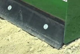 Close-up of the lower edge of a box blade attachment, showing a black steel wear blade secured with bolts, resting on sandy ground