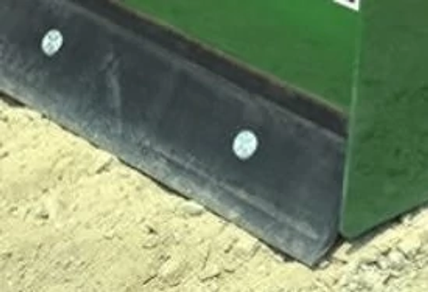 Close-up of the lower edge of a box blade attachment, showing a black steel wear blade secured with bolts, resting on sandy ground