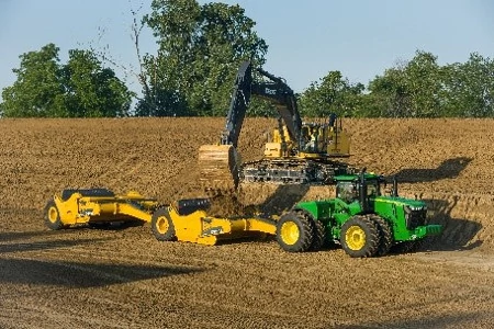 An excavator on dirt jobsite drops dirt into tandem, top-loading scrapers pulled behind a large green tractor