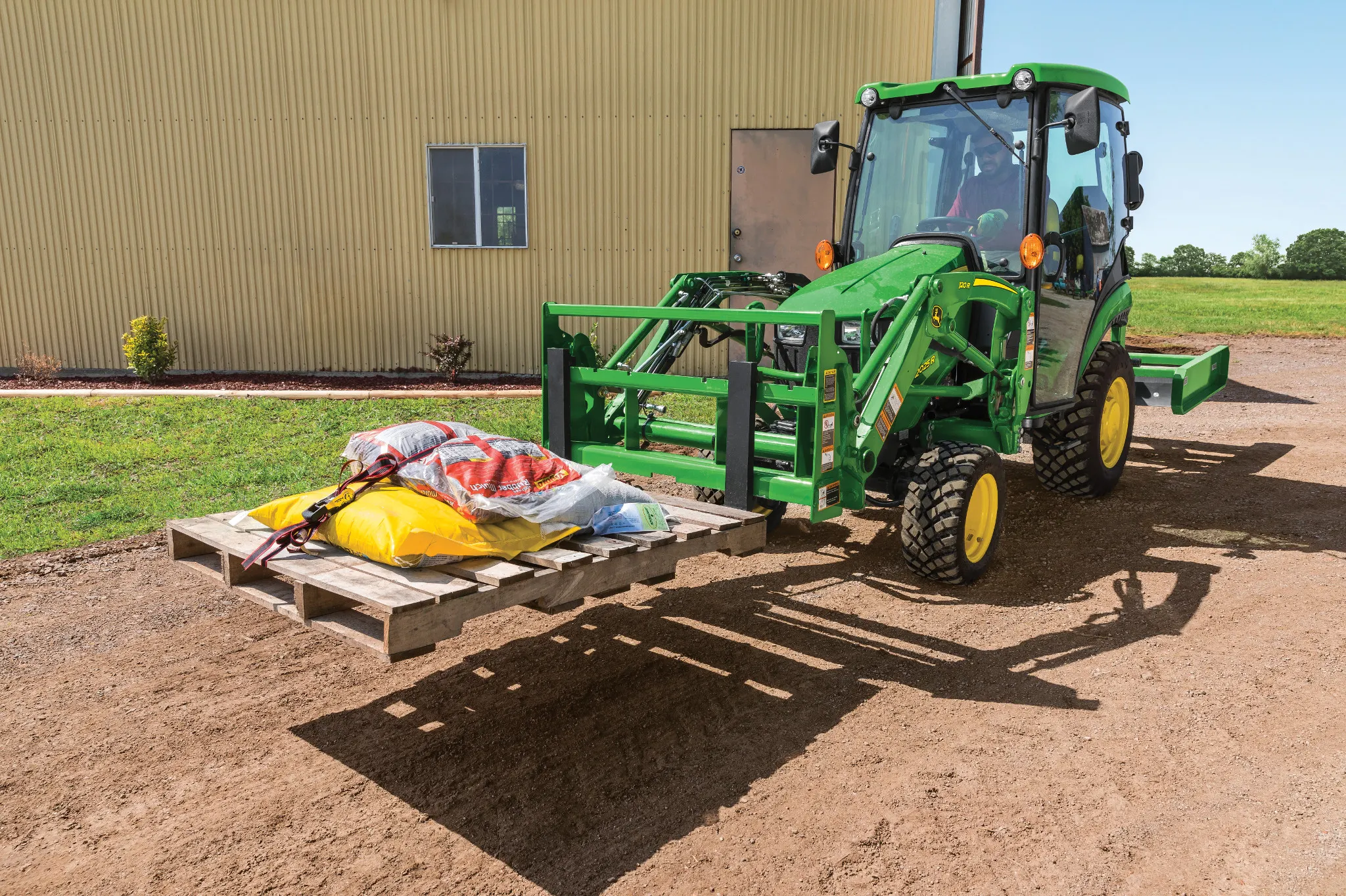 Green tractor with pallet forks lifts a loaded wooden pallet beside a metal building on a gravel surface, with open farmland visible in the background under clear sky