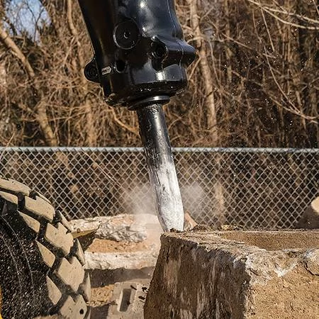 Close-up side view of a hydraulic hammer bit resting atop a concrete block on a dirt jobsite