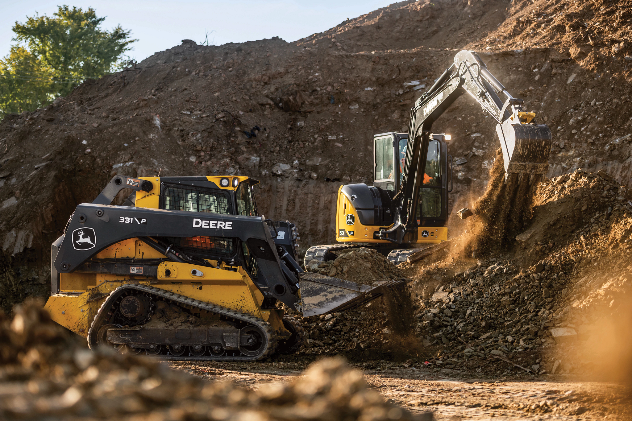 Tracked skid steer loader and compact excavator working together on a dirt hillside, with the excavator dumping soil and rocks into the loader’s bucket at a construction site