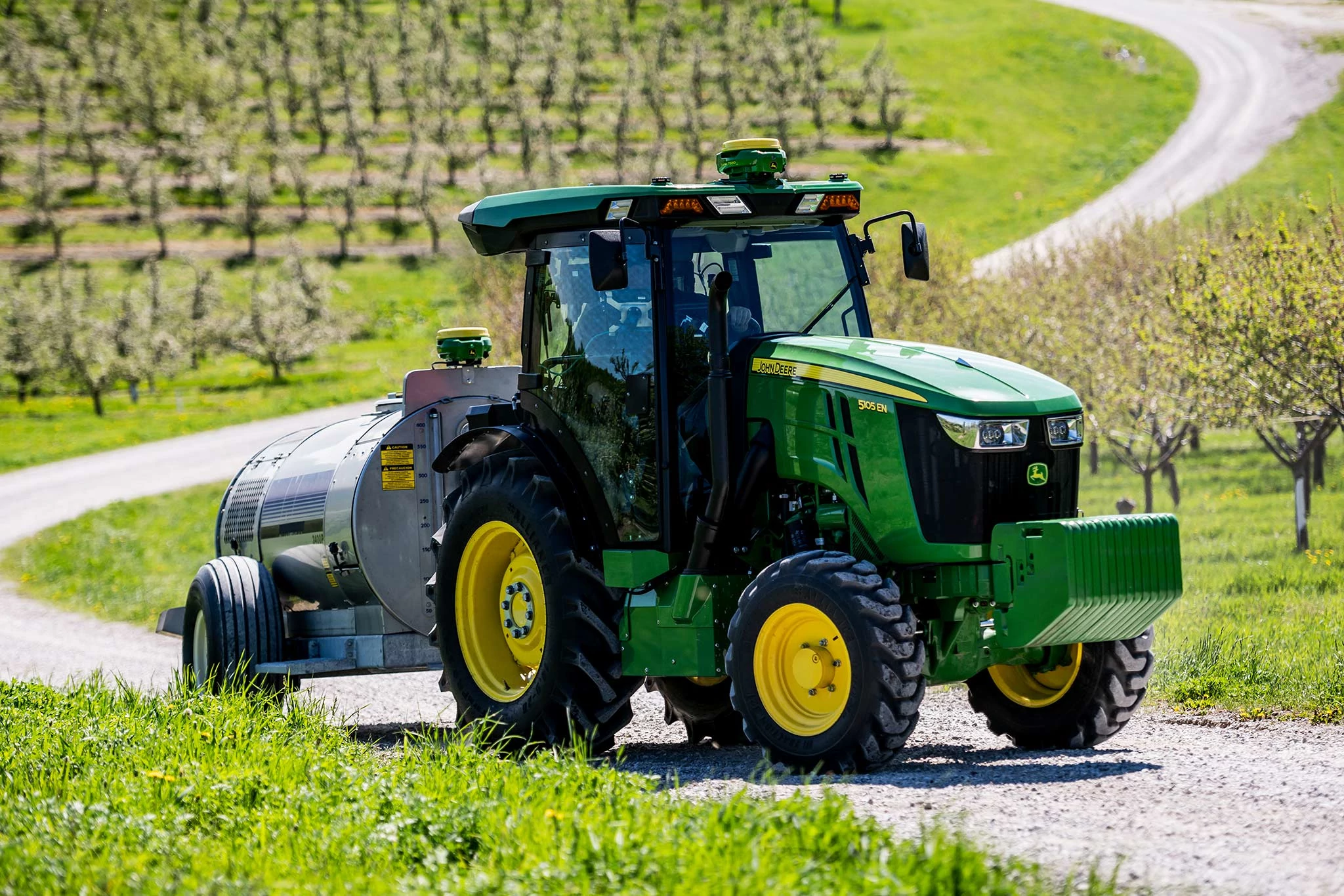Side right view of a 5105EN high value crop tractor towing a silver tank down a winding gravel road in an orchard