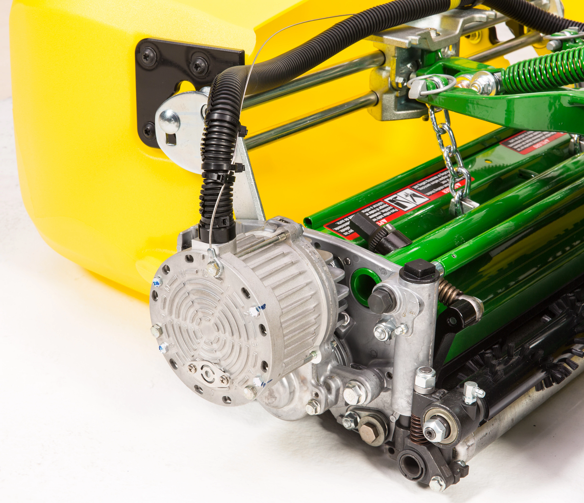 Close-up of mower attachment showing a silver electric motor and gearbox connected to rotating green rollers, with visible wiring, chain drive components, and mounting hardware against a white background