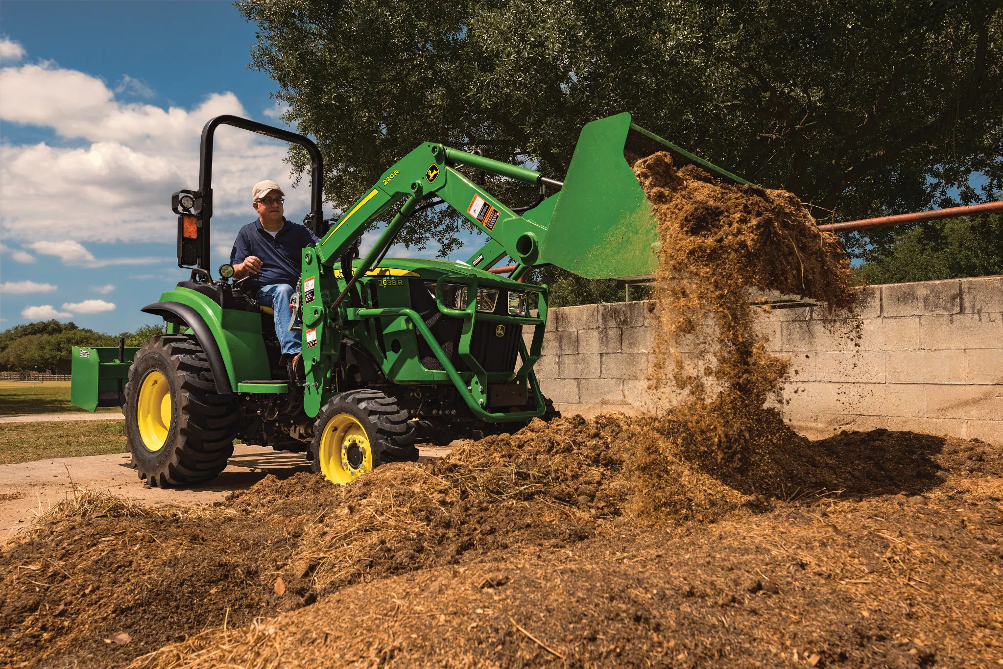 Green tractor with a front loader lifts and dumps a pile of fertilizer beside a concrete barrier, with loose soil scattered on the ground. Trees and a bright blue sky with clouds are visible in the background.