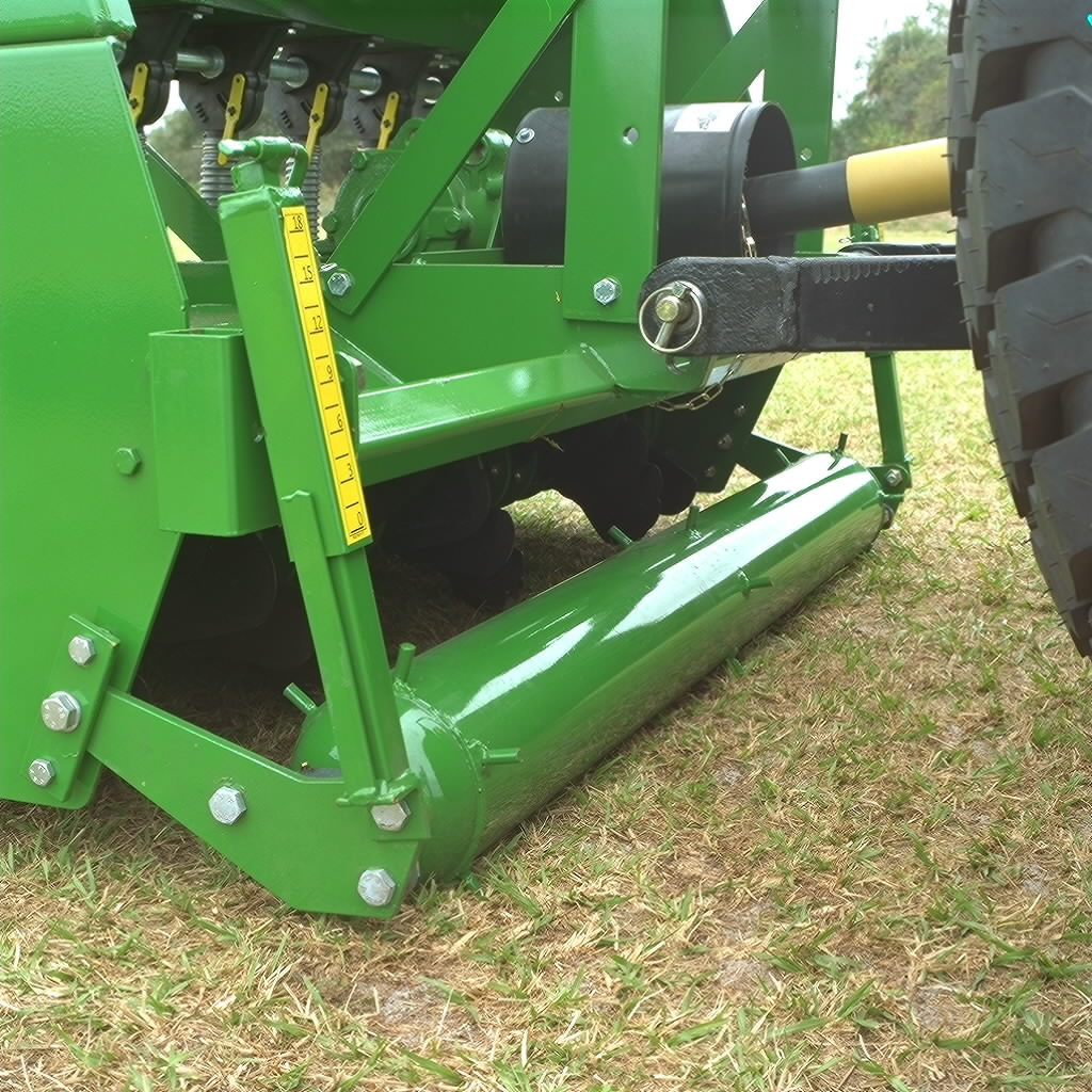 Close-up of a green overseeder implement showing a front roller with a yellow depth gauge and linkage arms above it, positioned on grass