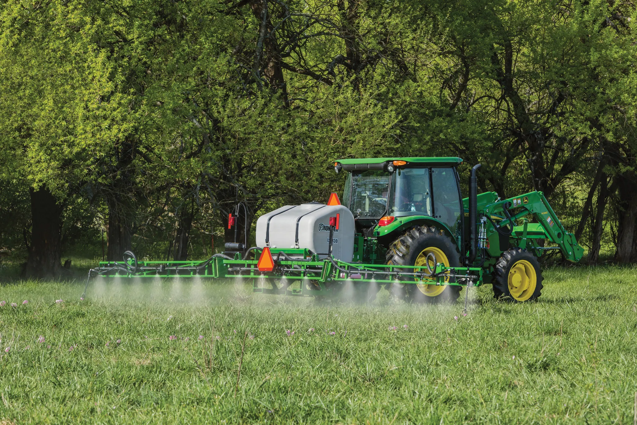 A tractor with a boom sprayer drives across a grassy field, spraying a fine mist over the crop, with leafy trees forming a dense green background.