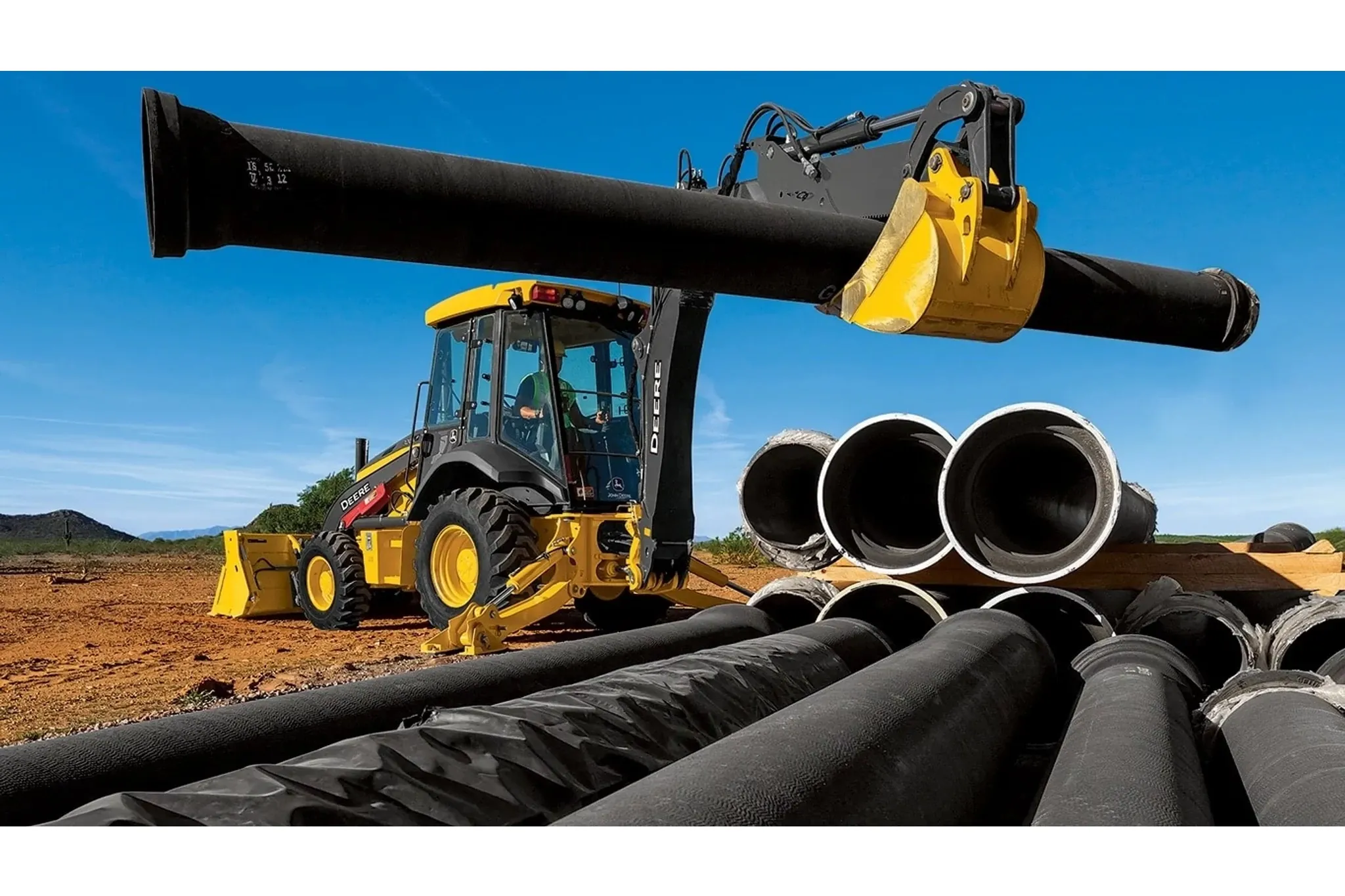 310 G-Tier Backhoe Loader carrying a piece of pipe to a pile for pipeline construction in large open field