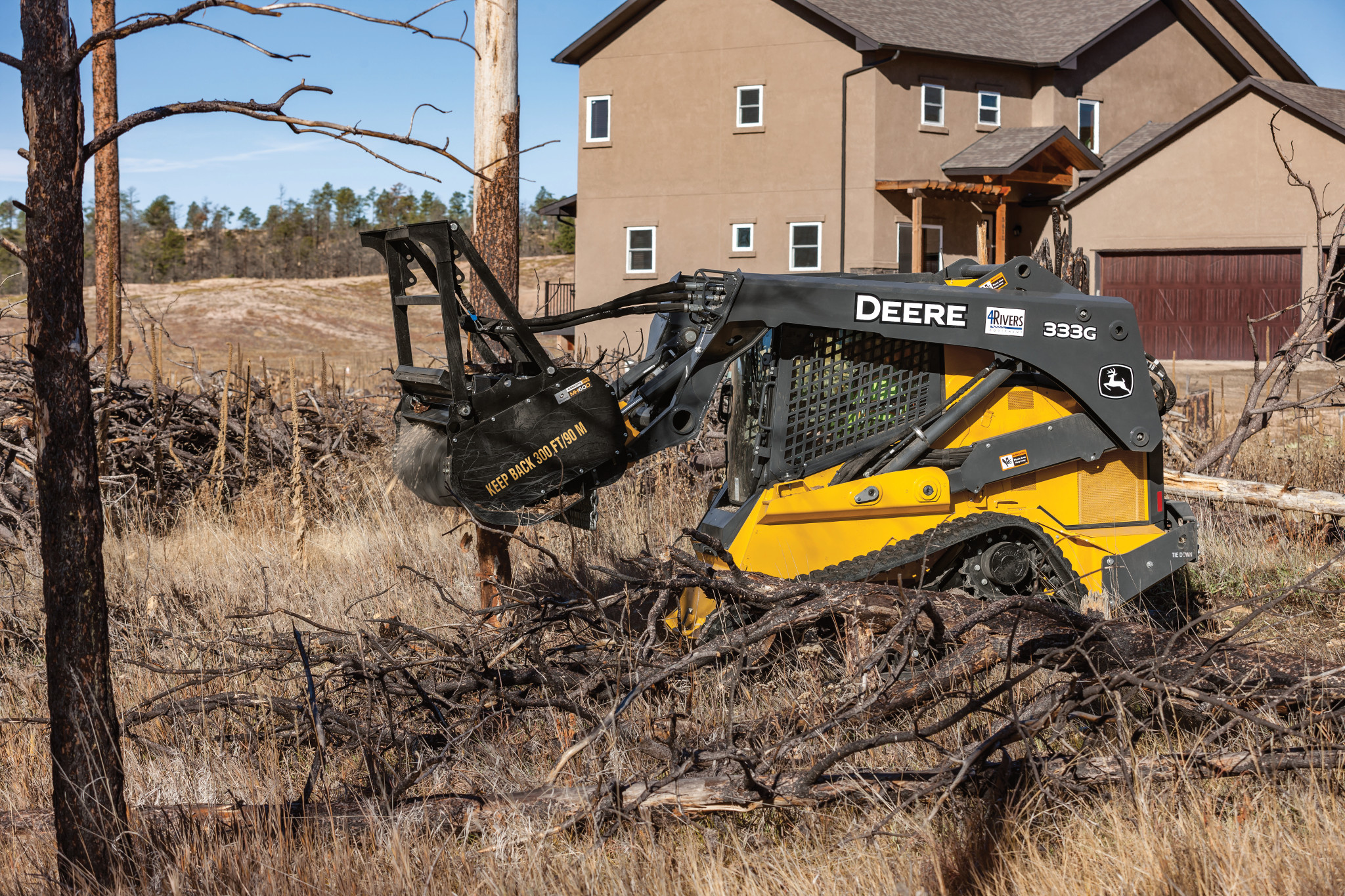 Skid steer loader using a front-mounted mulching head to grind branches in a dry, brushy area in front of a house
