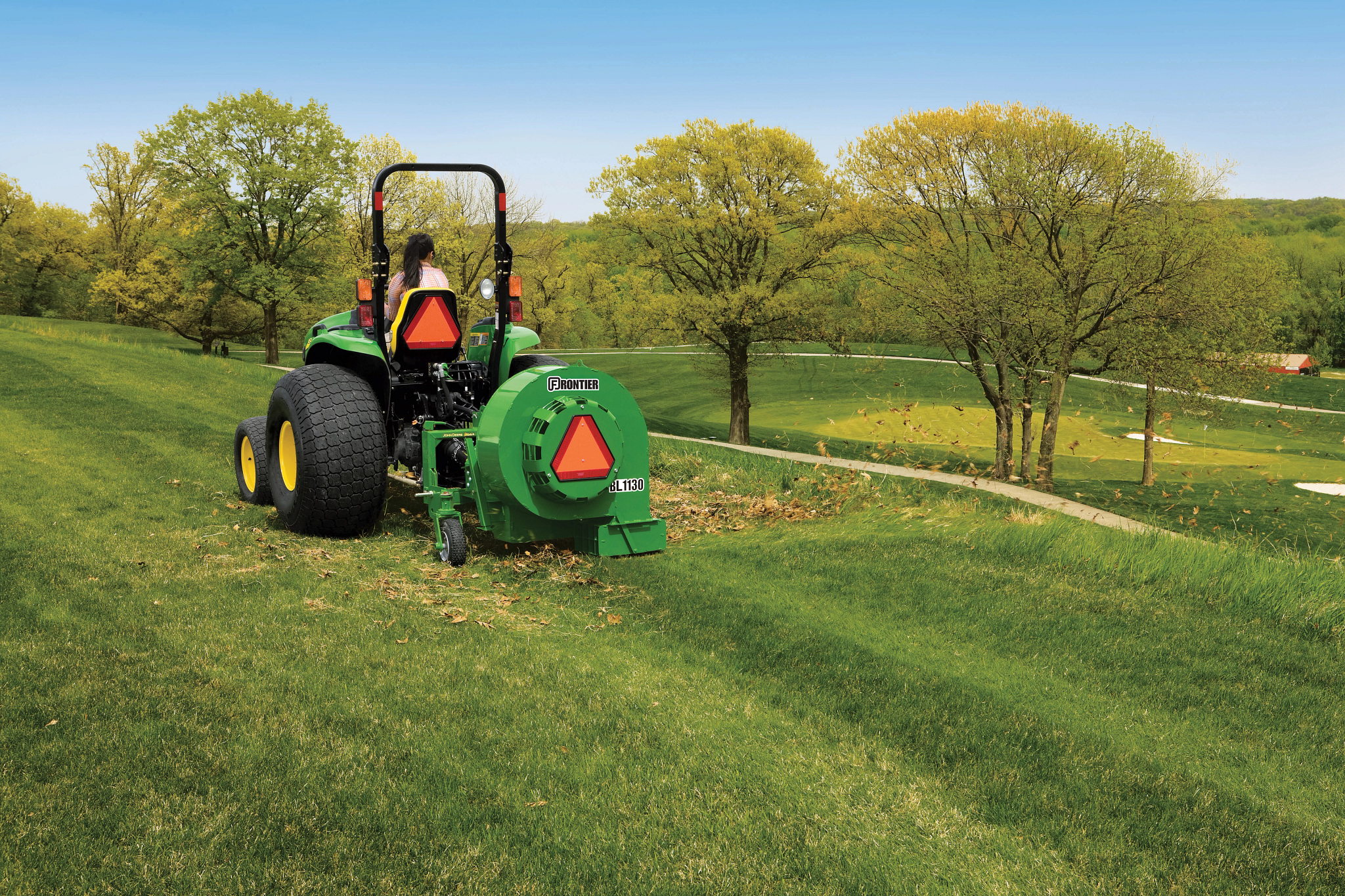 A green tractor operating on grass pulling a rear‑mounted blower attachment that is dispersing leaves with rolling hills, mature trees, and a golf course in the background