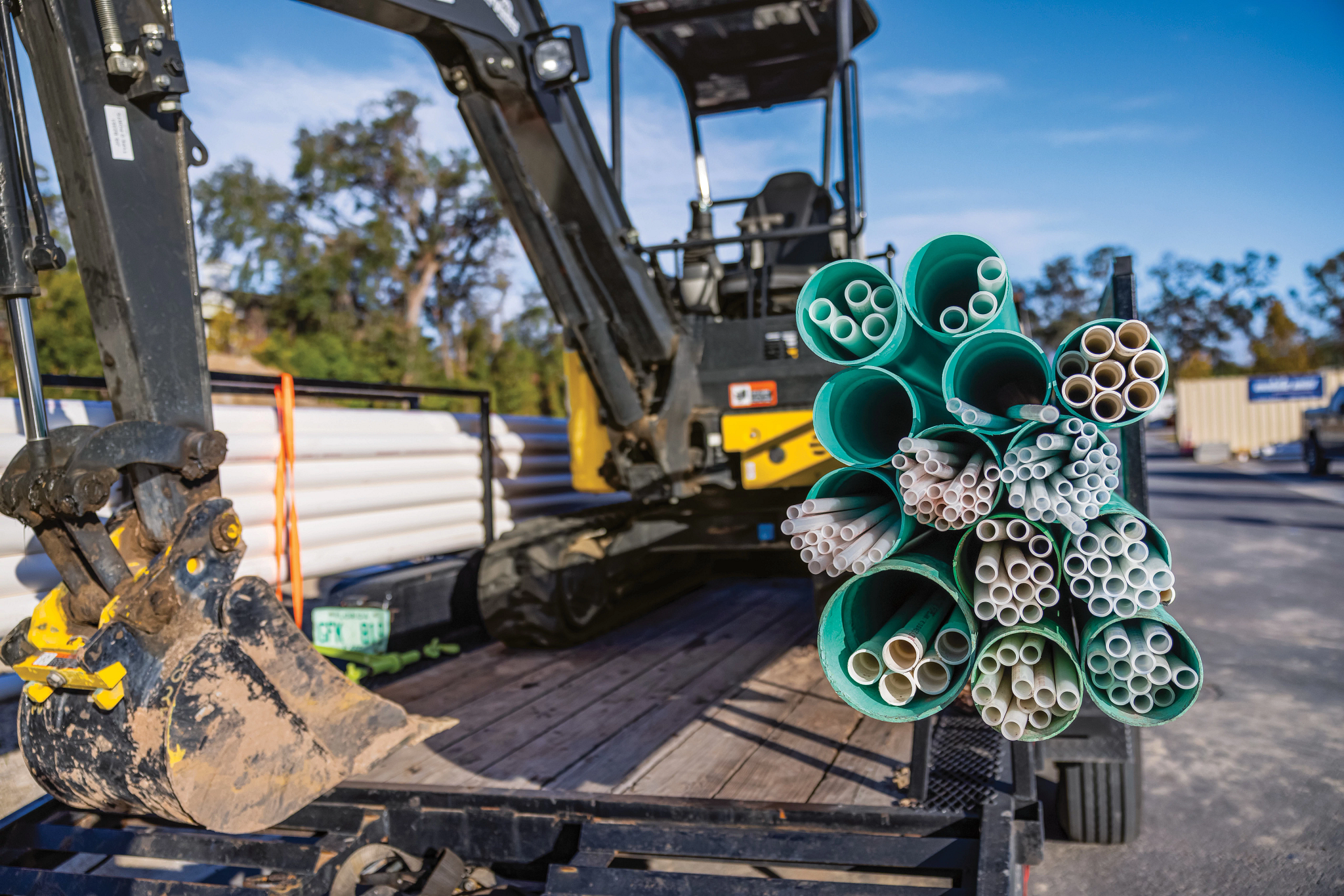 Close view of stacked green and gray plastic conduit pipes bundled on a trailer, with a yellow excavator and its dirt-stained bucket in the background at a construction site
