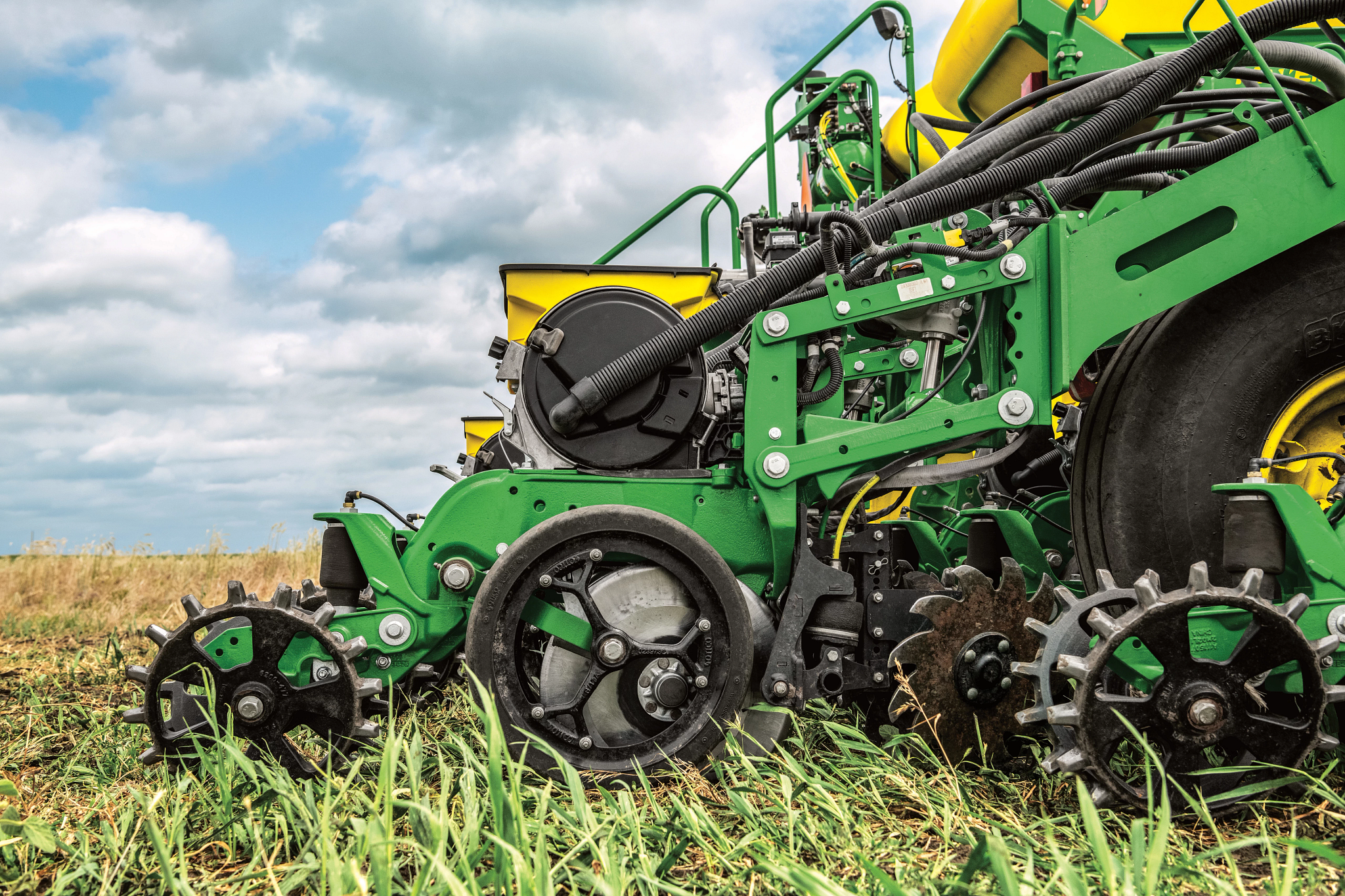 Close-up of a green farm planter in a field, showing metal wheels, hoses, and planting discs cutting through low crops. A yellow seed hopper is partially visible, with soil and cut vegetation beneath a cloudy sky.