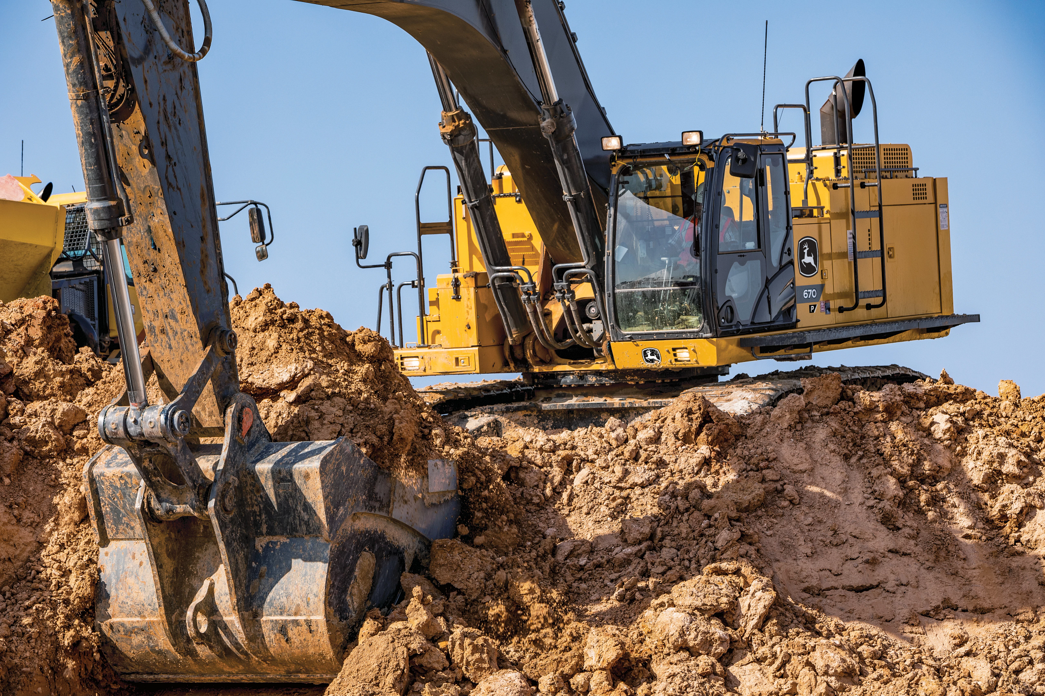 Close-up view of a yellow John Deere excavator working on a muddy construction site. The large metal bucket in the foreground is partially filled with wet soil as it digs into a thick pile of earth