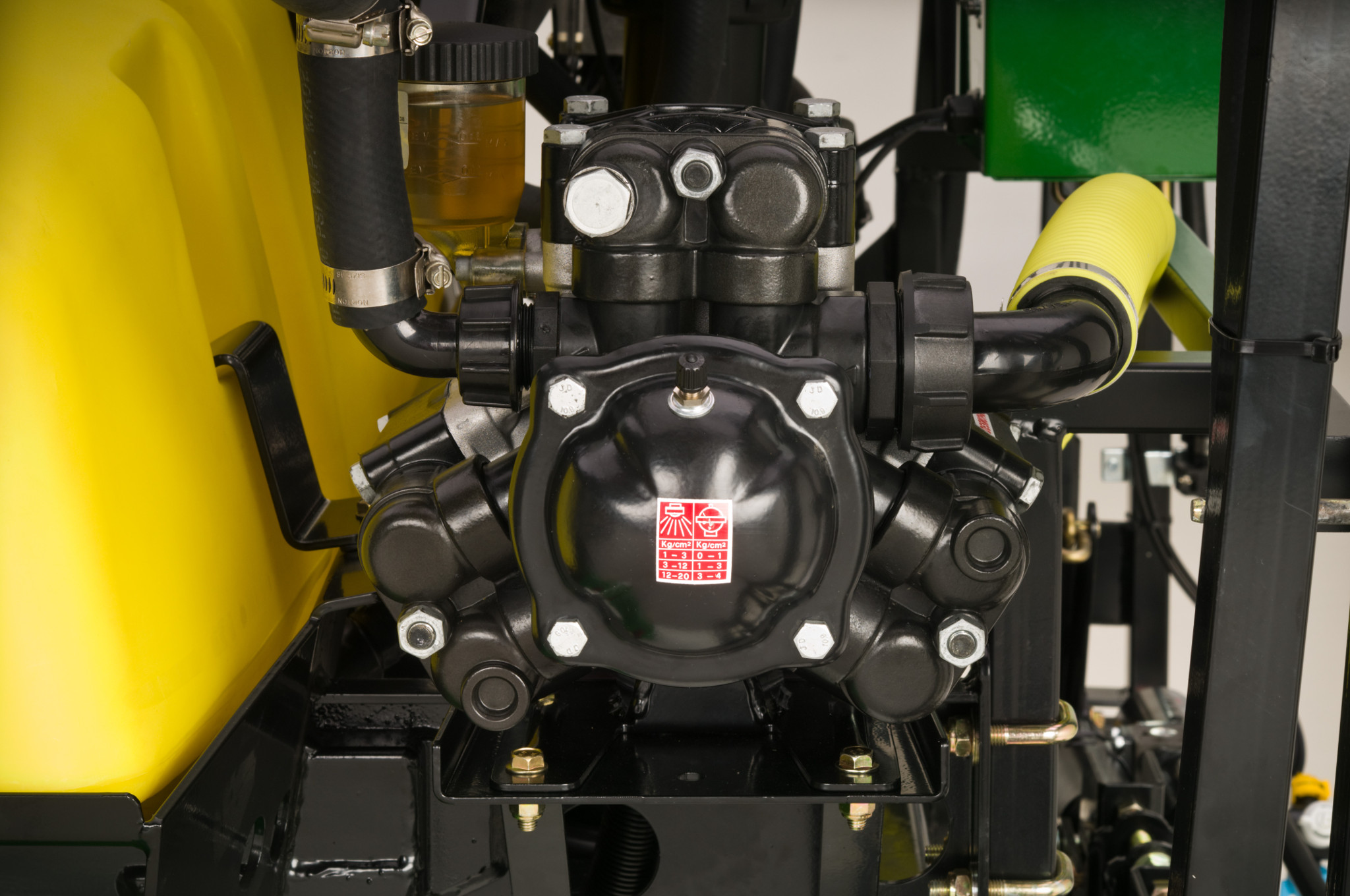 Close‑up view of a black hydraulic pump assembly with hoses, fittings, and mounting bolts attached to a yellow liquid tank inside agricultural equipment.