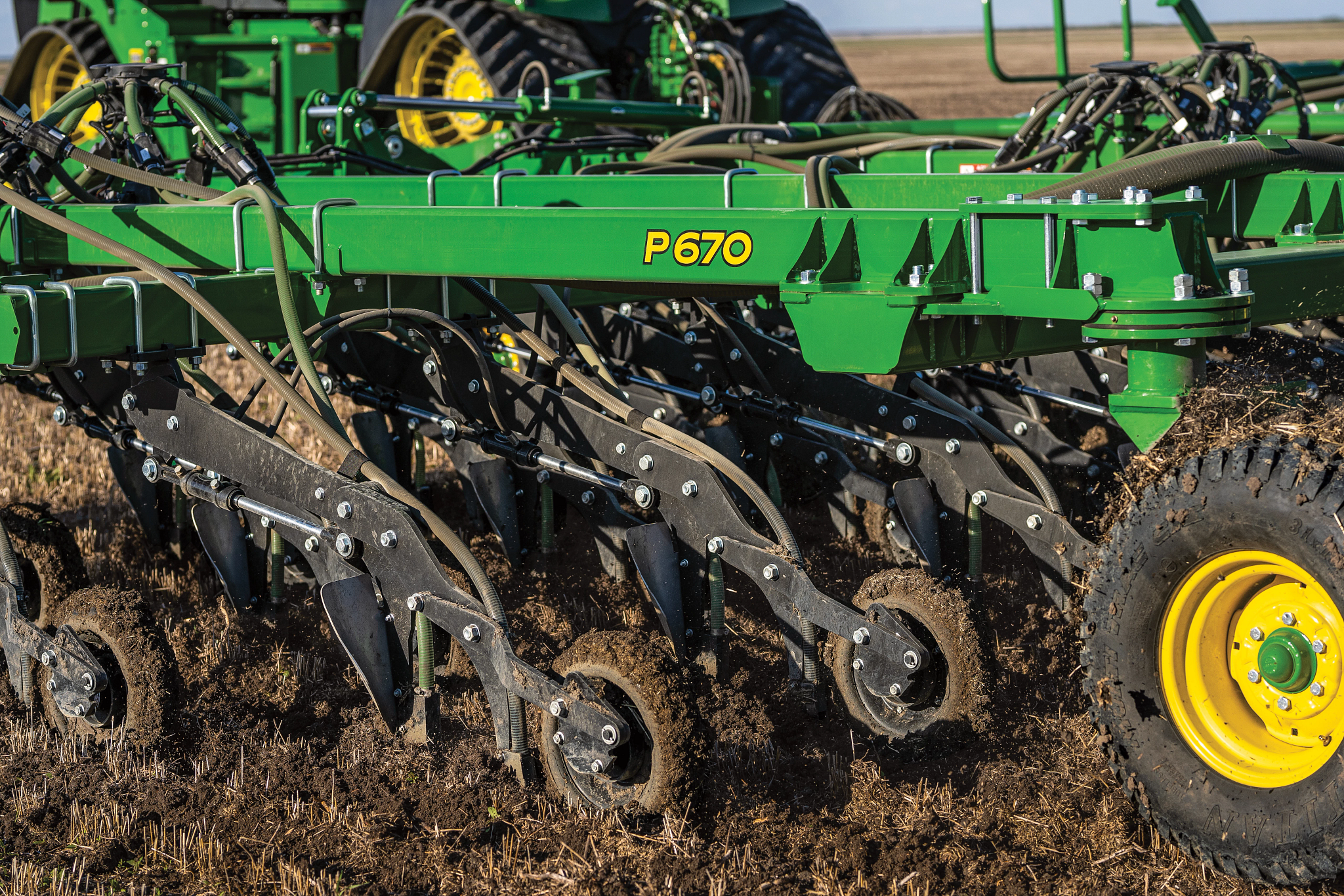 Green John Deere P670 Air Hoe Drill with multiple row units cutting through soil, showing angled discs, tines, and a yellow-rimmed wheel, operating in a field with crop residue visible on the ground