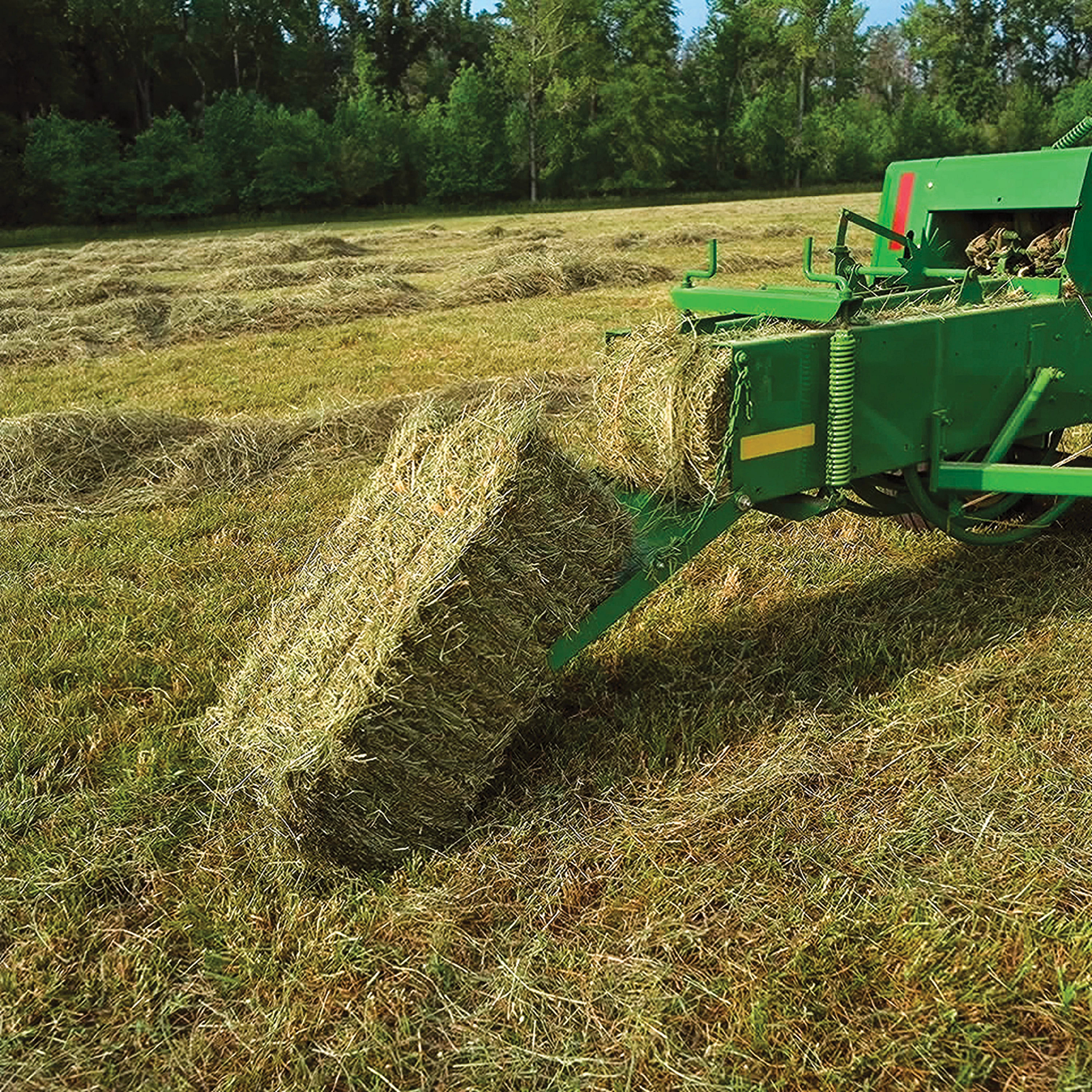 John Deere small square baler ejecting a finished rectangular hay bale onto a field, with windrowed hay visible behind the baler and trees lining the background.