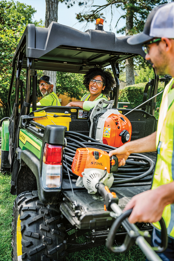 Two hi-vis-shirted operators are turning around in the seats of an XUV825M S4 4x4 Gator, laughing at a third person unloading a weedwhacker from the cargo box.